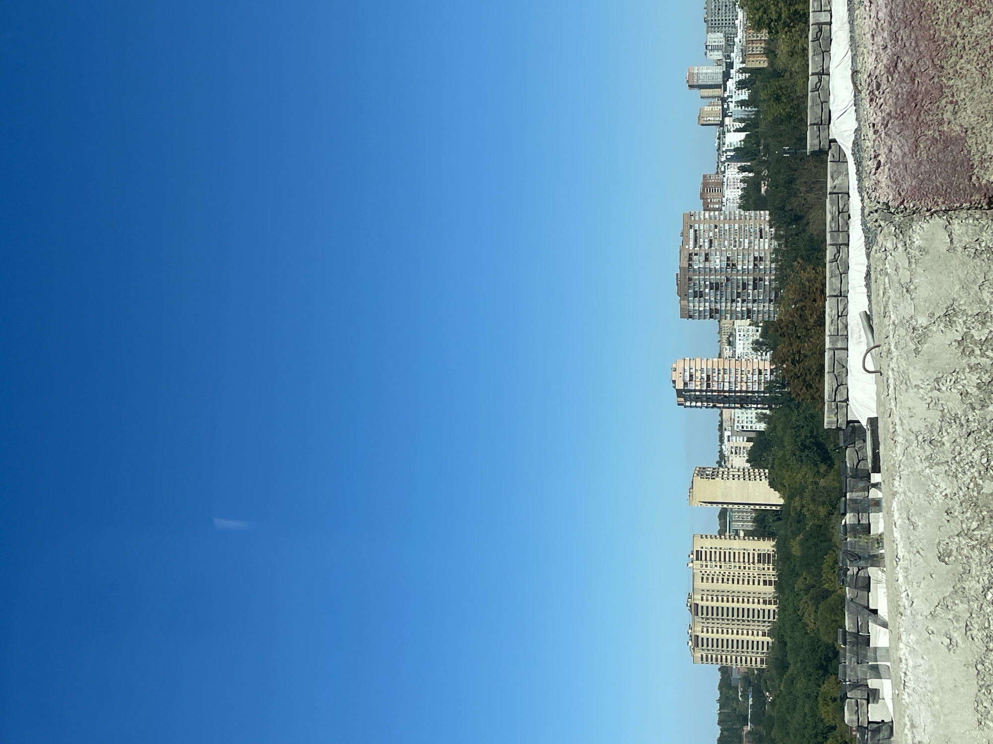 This is an outdoor, high-angle, eye-level, medium shot photograph of a cityscape under a clear blue sky. In the foreground, a rough, textured concrete structure with visible aggregate and peeling paint spans the lower portion of the frame. This structure appears to be the edge of a rooftop or elevated platform.
Beyond the foreground structure, a line of trees forms a dense green band across the middle ground of the image. Behind the trees, a collection of urban buildings rises, predominantly apartment complexes of varying heights and architectural styles. Some buildings are taller and more modern, while others are shorter and appear older.
A thin, vertical smudge, possibly a faint contrail from an aircraft, is visible in the upper portion of the otherwise clear blue sky. The lighting suggests it is daytime, with the sun illuminating the scene from the right. There are no people or animals visible in the frame. The scene depicts a typical urban residential area, likely in Rostov-on-Don, Russia, given the architectural style of the apartment buildings and the presence of mature trees.