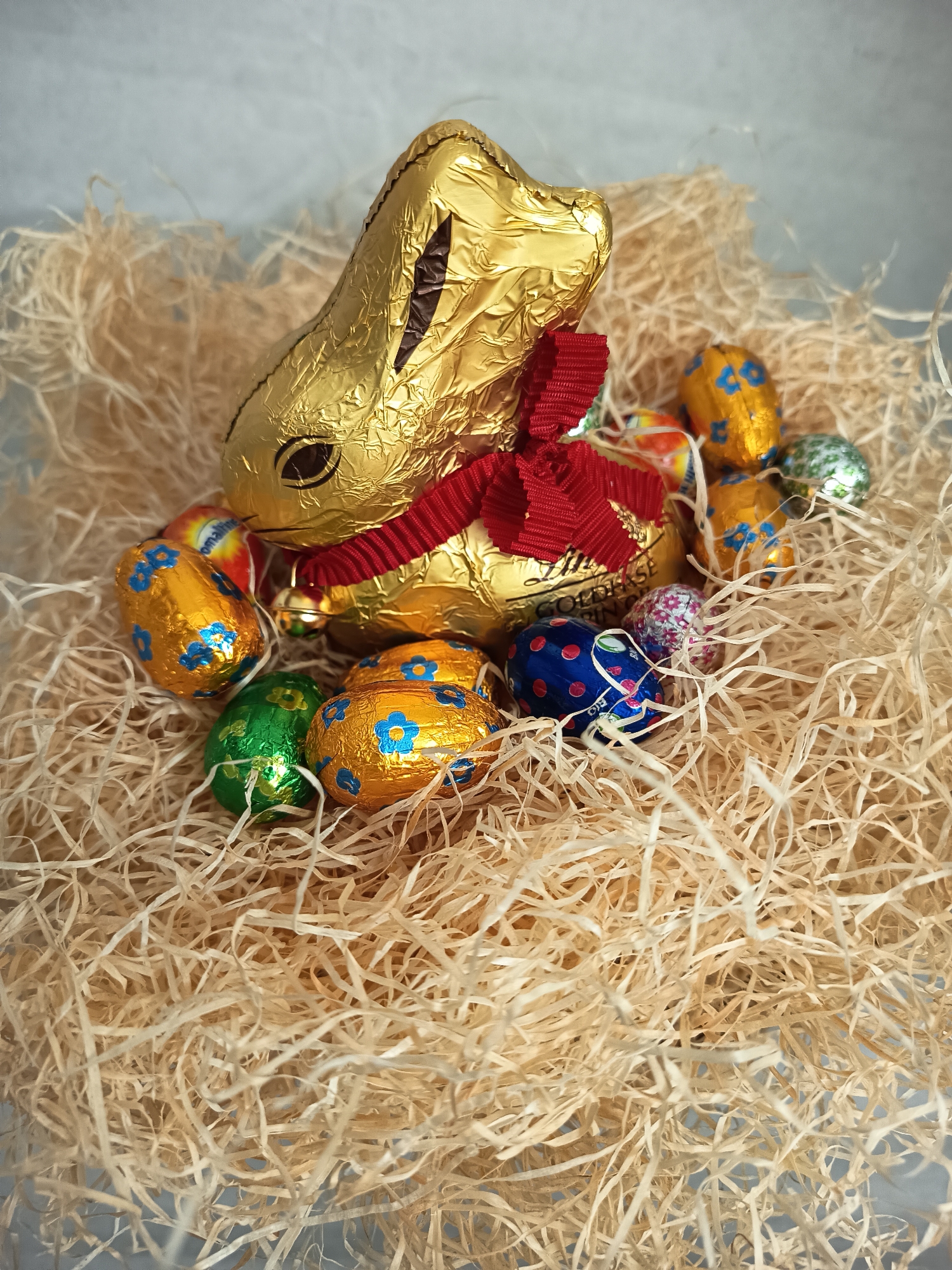 A photograph depicts a gold foil-wrapped chocolate bunny, adorned with a red ribbon, surrounded by numerous small, foil-wrapped chocolate eggs, arranged within a bed of wood shavings. The eggs are wrapped in various colors and patterns, including gold with blue flower designs, green, silver, and blue with red polka dots. The bunny is positioned towards the upper-center of the image, and the composition is filled with the texture of the wood shavings. The chocolates appear to be Lindt products, which are commonly found in Wangen bei Olten, Switzerland.