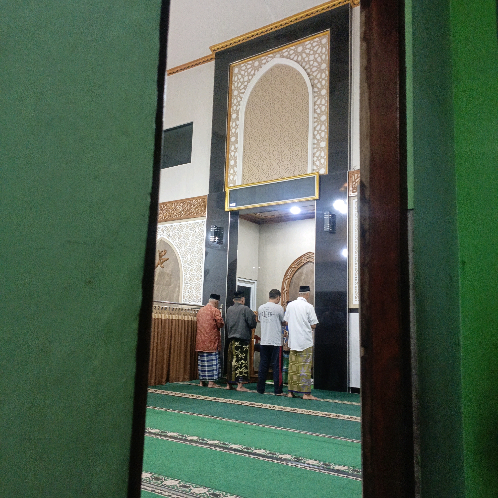 The image depicts the interior of a mosque, possibly in Salatiga, Indonesia, as indicated by the context. Four men are standing on a green carpet with prayer mat markings, facing an ornate wall. The men are dressed in traditional Islamic attire, including sarongs and caps. The wall behind them features intricate Islamic geometric patterns and calligraphy, with a large arched motif in the center. A brown curtain hangs along the left side of the wall. The lighting appears to be artificial, with lights visible on the wall. The perspective suggests the photo is taken from an entrance or doorway, with a green wall element on the left and a dark wooden frame on the right partially obscuring the view.