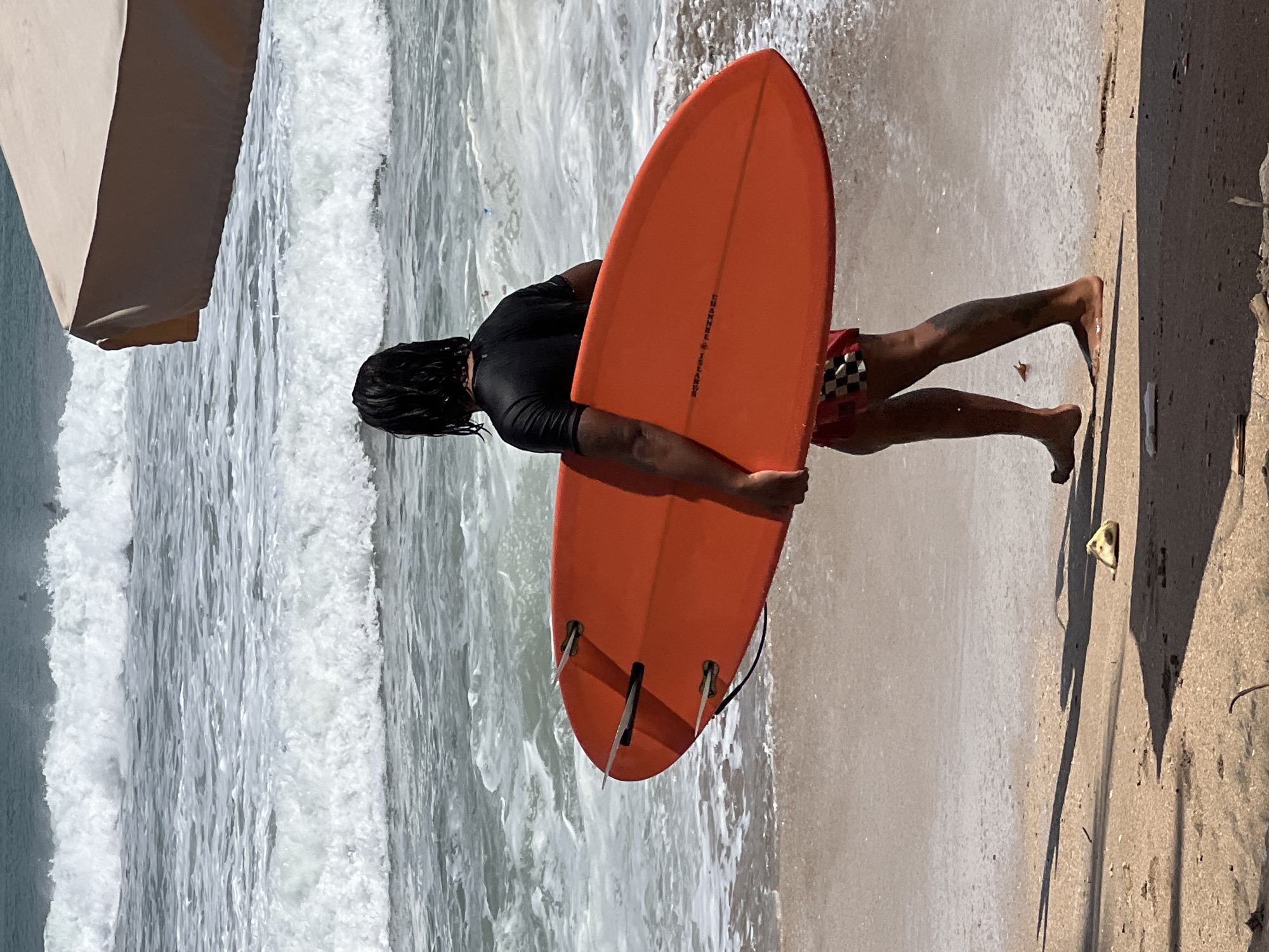 A person, viewed from behind, walks on the sand at the edge of the ocean, carrying an orange surfboard. The person has dark, wet hair, is wearing a black t-shirt and red and black checkered swim shorts. They have tattoos on their legs. The surfboard has three fins visible on the bottom. The ocean has waves breaking on the shore, and a portion of a tan umbrella is visible in the upper right corner of the image. The scene appears to be at a beach in Kuta, Indonesia, given the characteristics of the waves and the presence of surfboards.