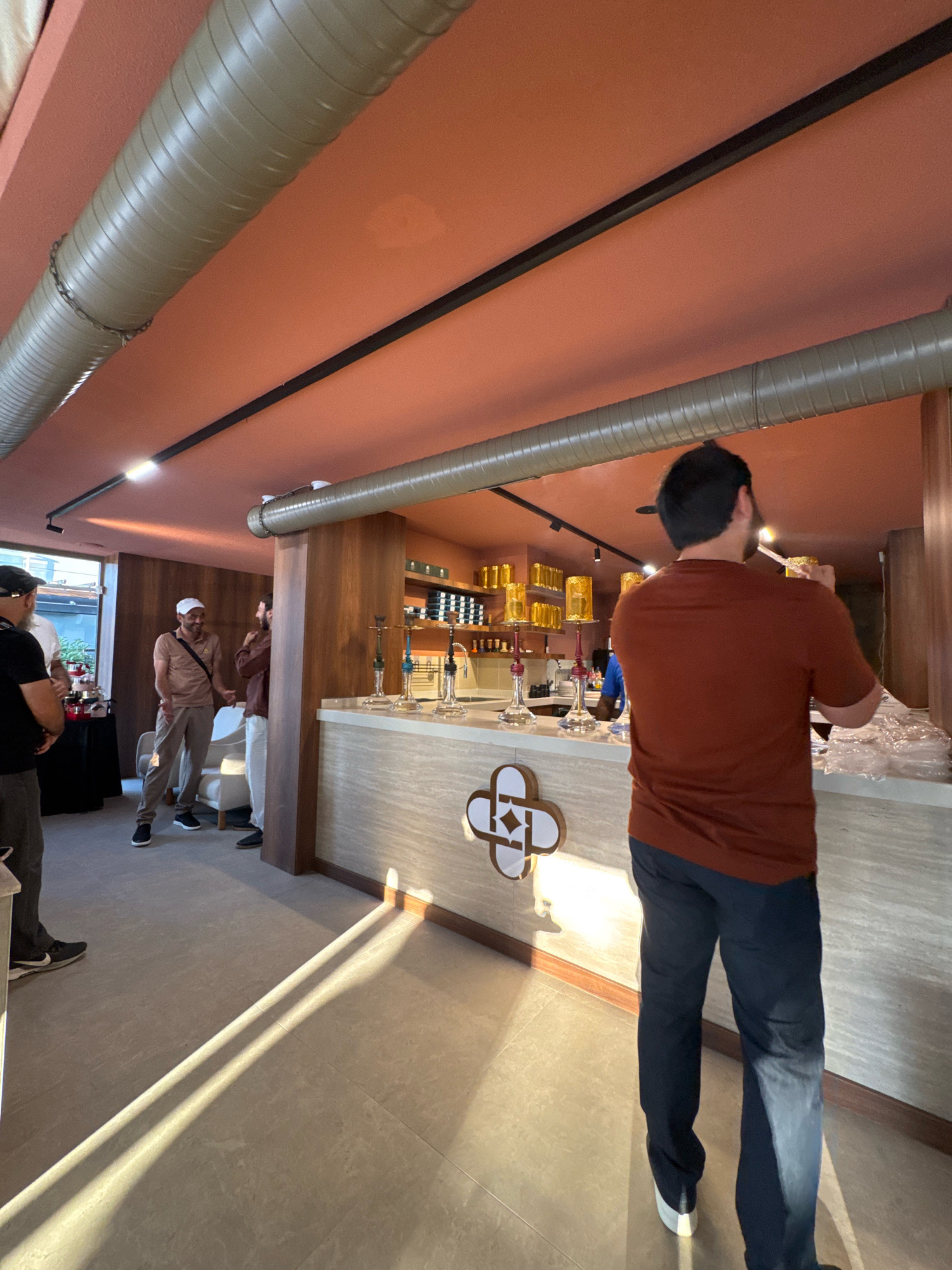 The image depicts an indoor scene, likely a shop or establishment in Antalya, Turkey. In the foreground, a person wearing a reddish-brown shirt and dark pants is seen from behind, holding a glass pipe to their face, suggesting they are smoking a hookah. The person is standing at a counter made of light-colored stone with a geometric pattern embedded in it. Behind the counter, several hookahs with decorative bases are displayed, along with shelves stocked with bottles and other items. The ceiling is a muted orange color with exposed, ribbed, olive-green ductwork.
To the left of the counter, three men are visible. One man, wearing a light brown shirt and pants and a white cap, is facing another man in a brown shirt and white pants, and they appear to be in conversation. Another man, wearing a black t-shirt and a black cap, is standing partially visible on the far left. In the background on the left, there is a black table draped with a black cloth and some red items, possibly merchandise. Sunlight is casting strong diagonal shadows across the tiled floor. The overall impression is of a modern, well-lit retail space with a focus on hookah products.