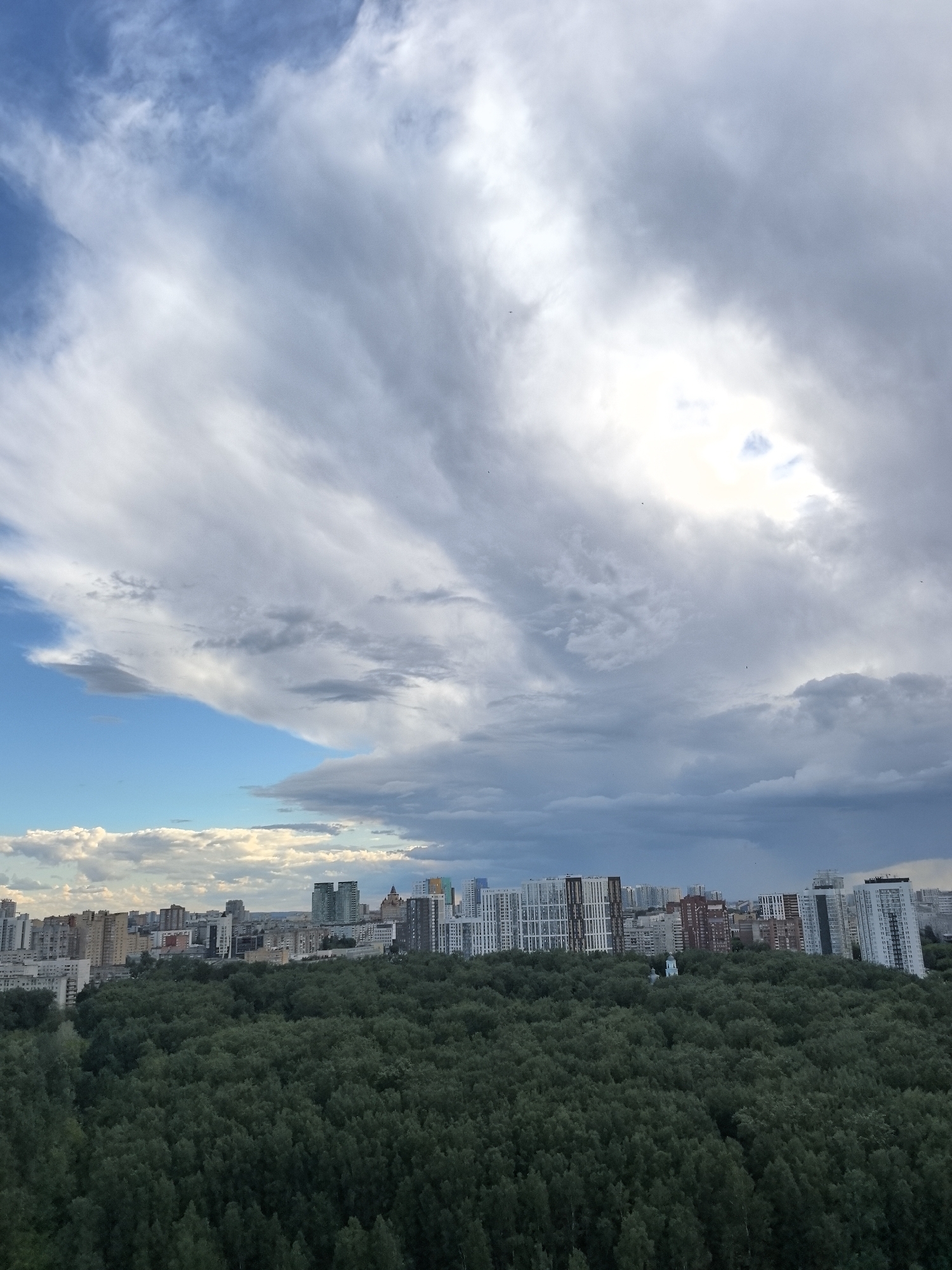 The image presents an elevated view of Perm, Russia, showcasing a dense urban forest in the foreground. Behind the forest, a cityscape emerges, with various high-rise buildings and residential structures visible against the skyline. The sky dominates the upper portion of the frame, displaying a mix of dynamic cloud formations, ranging from light, wispy clouds to darker, more ominous storm clouds. A patch of clear blue sky peeks through the clouds on the left side of the image. A small, light-colored structure resembling a church or temple is partially visible amongst the trees, providing a focal point within the forest canopy. No people are visible in the image.