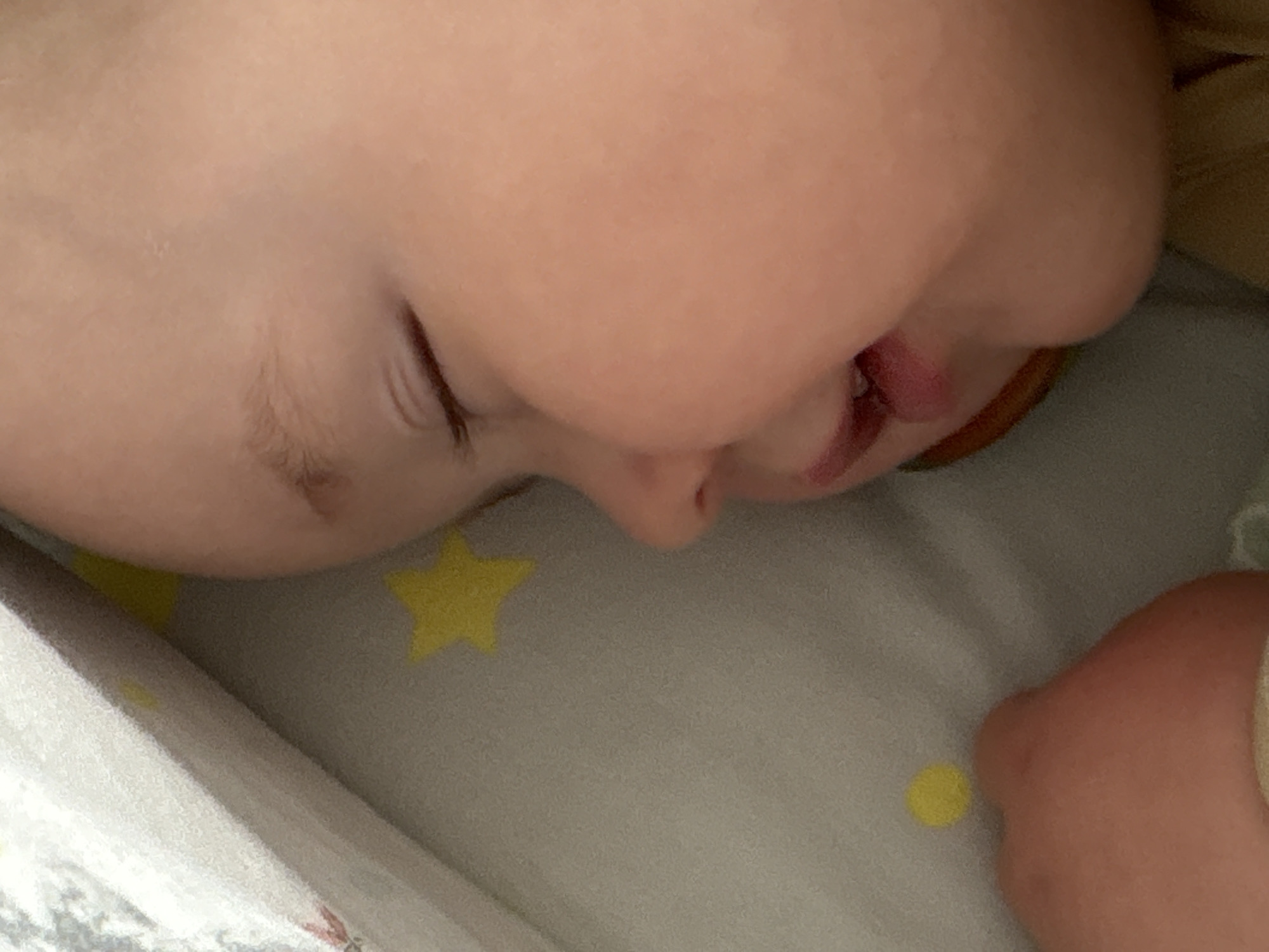 A close-up photograph shows a baby sleeping on a patterned grey and white blanket adorned with yellow stars. The baby is facing to the side with its eyes closed. The baby's skin is light-toned. A corner of the baby's mouth is slightly open. The setting is indoors. The location of the image is unknown and there is no indication it was taken in Izhevsk, Russia.