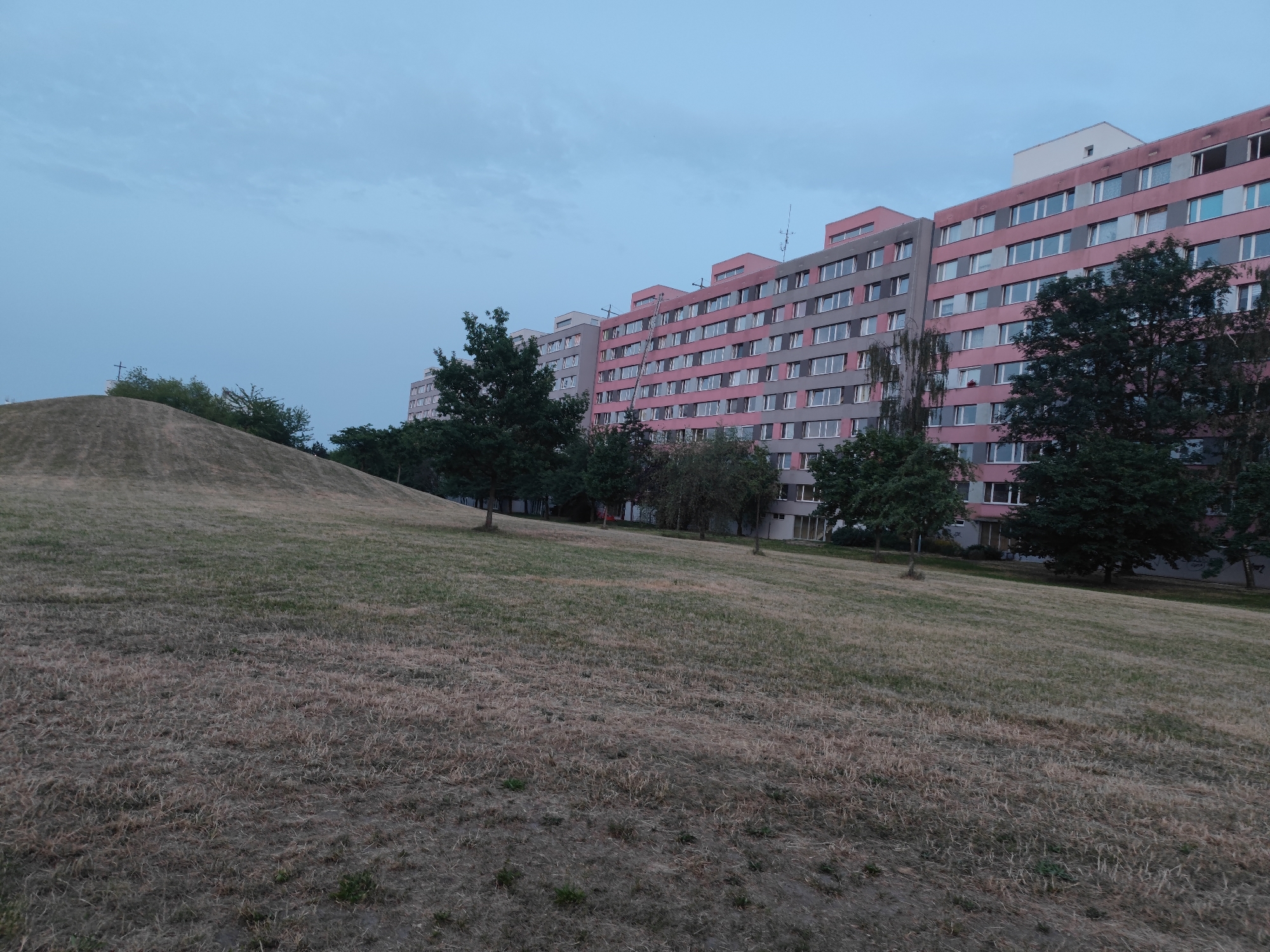 The image depicts a residential apartment building in Prague, Czechia. The building is long and has multiple floors, with a facade predominantly in shades of pink and grey. Numerous windows line the building. In front of the building, there is a large, grassy hill that appears somewhat dry and mowed. Several trees are situated between the hill and the building. The sky above is overcast and a pale blue. The scene appears to be captured at dusk or dawn.