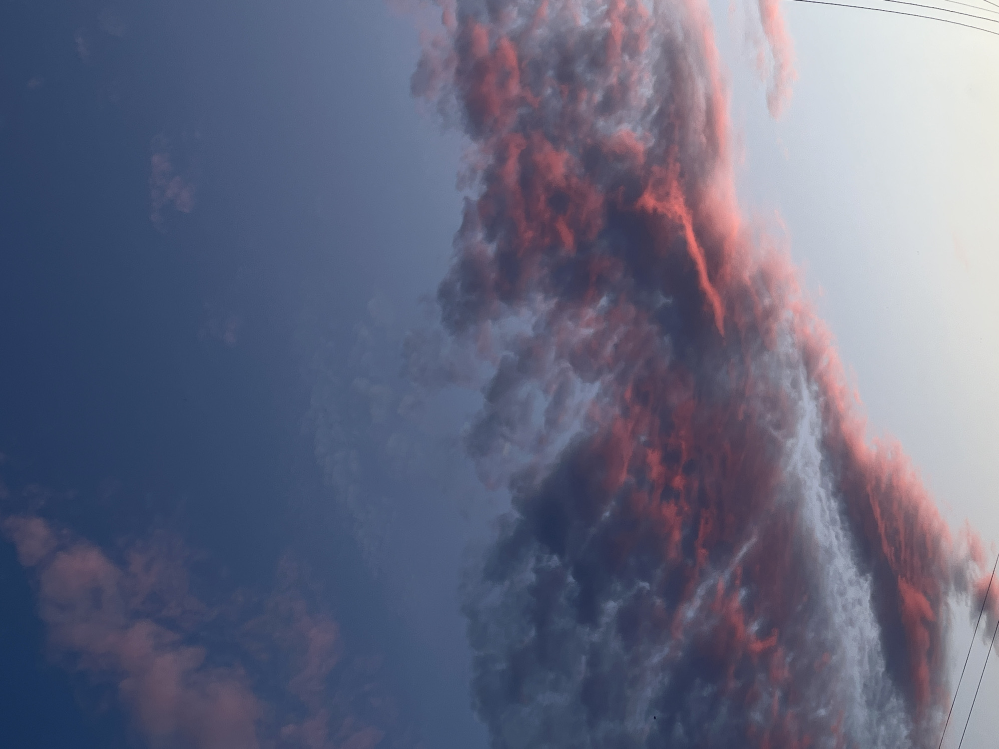 The image shows a sky with a mix of blue and light grey hues. A large cluster of clouds with pink and reddish tones dominates the right side of the frame. Two overhead power lines can be seen in the upper right corner. Based on the location provided (Фоки, Russia), the image is likely taken in or near this Russian city.