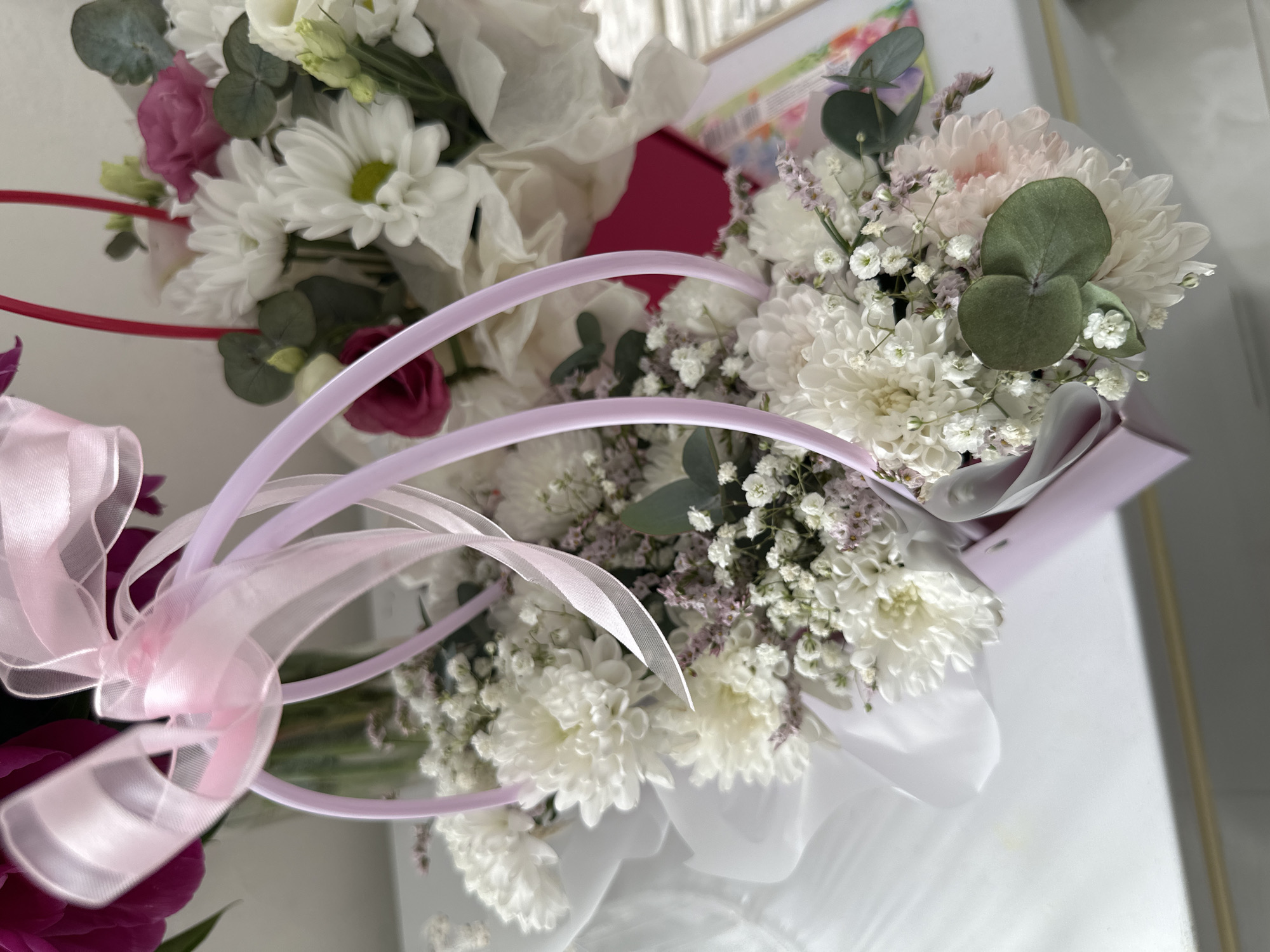 A still life photograph showcases two floral arrangements presented in brightly colored, rectangular gift boxes. The arrangement in the foreground, positioned to the left, is a bouquet of white chrysanthemums and delicate white and pink filler flowers, accented with green eucalyptus leaves. It is adorned with a pink ribbon and has pale lavender handles. To the right, a second, less distinct floral arrangement in a pink box also contains white flowers and greenery. Both arrangements appear to be placed on a white surface, possibly a tabletop or counter, with a hint of a colorful patterned object in the background. The lighting suggests an indoor environment.