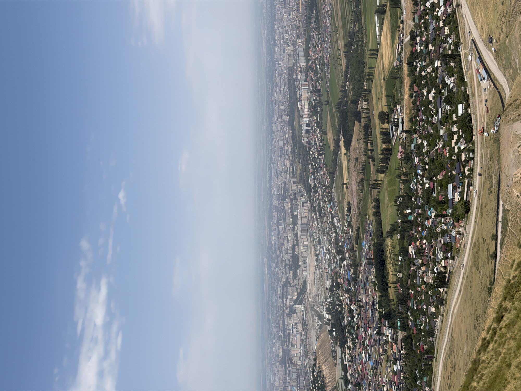 An aerial panoramic view shows a sprawling city with numerous buildings under a hazy sky. The foreground features a steep, grassy slope. In the midground, a densely populated residential area with houses featuring colorful roofs is visible. A road runs through this area, with several vehicles, including a truck and what appears to be construction equipment, present. Beyond the residential zone, the city expands with many apartment buildings and other structures stretching towards the horizon, where a faint line of mountains or hills is discernible. The overall atmosphere is one of urban expansion into a semi-arid landscape.