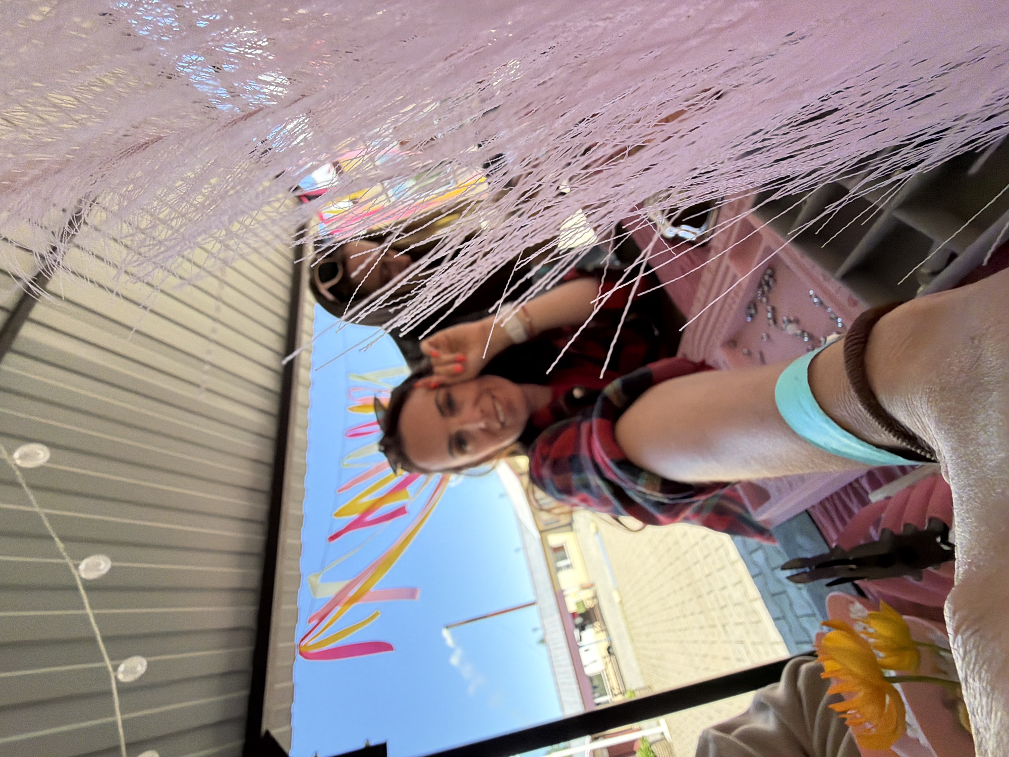 This is a selfie-style, medium shot of two women standing under a corrugated metal awning, outdoors. The woman in the foreground is holding the camera, and her left arm is extended towards the lens. She is wearing a red and black plaid shirt, a blue wristband, and a brown bracelet. Her right hand is raised, with fingers extended. She is smiling and looking at the camera. Behind her, partially obscured by a pink, stringy decoration, is another woman wearing sunglasses on her head and a black hoodie. She appears to be looking down. In the background, the sky is blue with scattered clouds. There are colorful ribbons hanging from the awning, and a building with a white roof and yellow walls is visible in the distance. The ground appears to be paved with interlocking bricks. There is a table or display area in front of the women, with some small orange flowers in a pink dish, and what appears to be jewelry or craft supplies. The overall scene suggests an outdoor market or festival.
