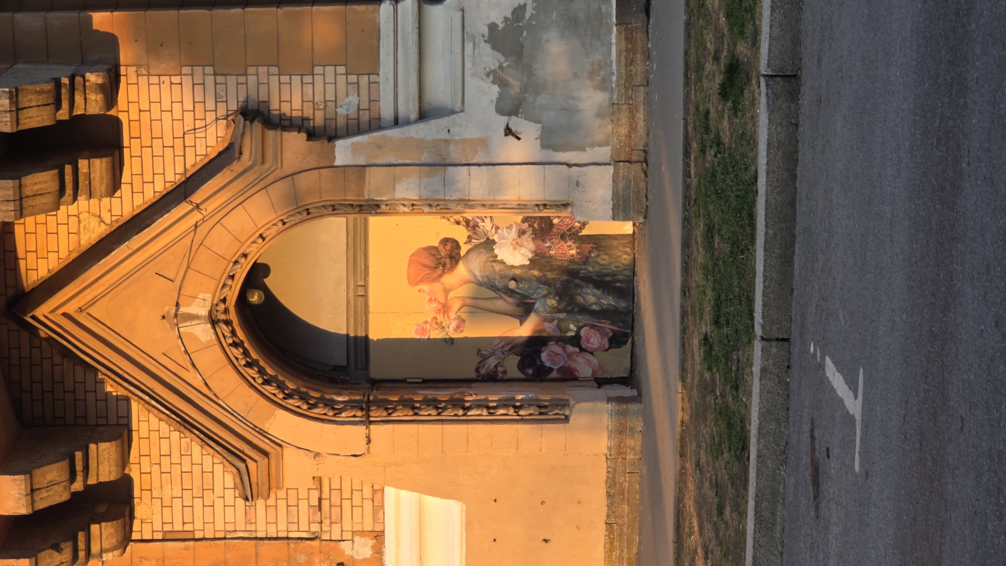 The image depicts a mural of a woman smelling flowers, painted on the wall within an arched doorway of a building in Saint Petersburg, Russia. The building is constructed of yellow-orange brick, with decorative molding around the archway. The mural faces a road, with grass and pavement visible to the right. The lighting suggests it is sunset or sunrise.