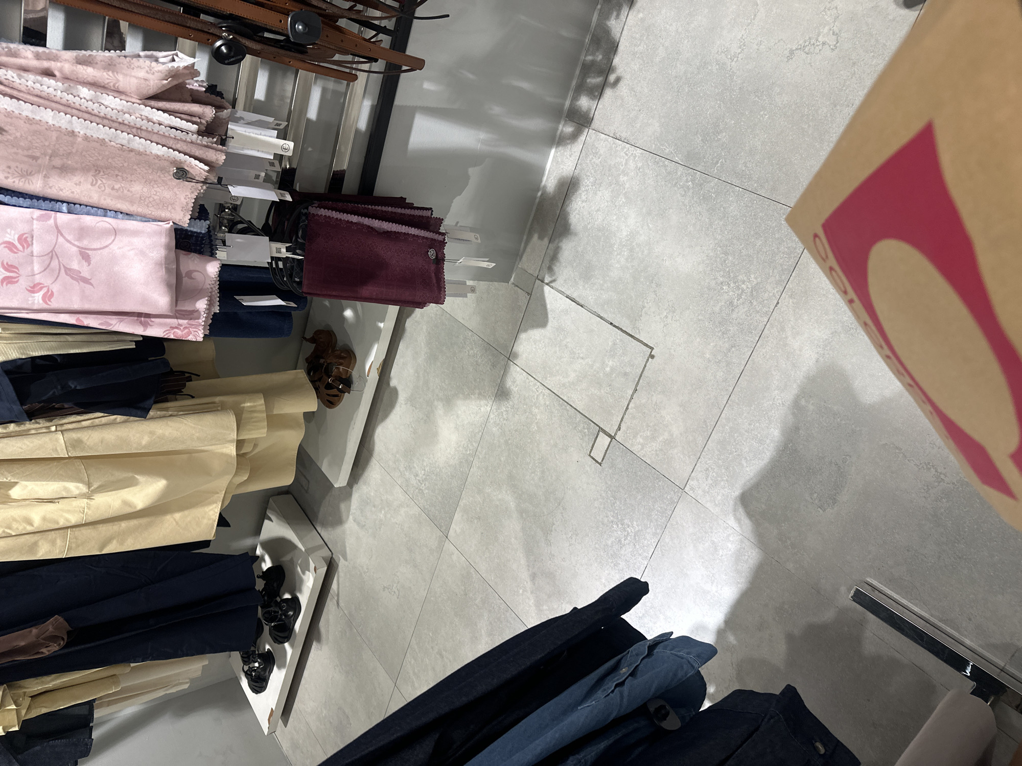 The image depicts the interior of a retail store, possibly a clothing boutique, with garments and accessories displayed. In the foreground, a person's hand holds a brown paper bag with a pink graphic design, suggesting a purchase. The floor is tiled with large, light gray squares. Garments, including what appear to be dresses or long skirts in beige and navy blue, are hanging from a rack on the left side. A selection of fabric swatches or scarves in shades of pink and burgundy are displayed on hangers in the middle and right sections of the image. Brown leather sandals are visible on a raised white platform near the navy blue garments. There are no identifiable landmarks or characteristics that would specifically place this scene in Denpasar, Indonesia, as the store's interior appears generic.