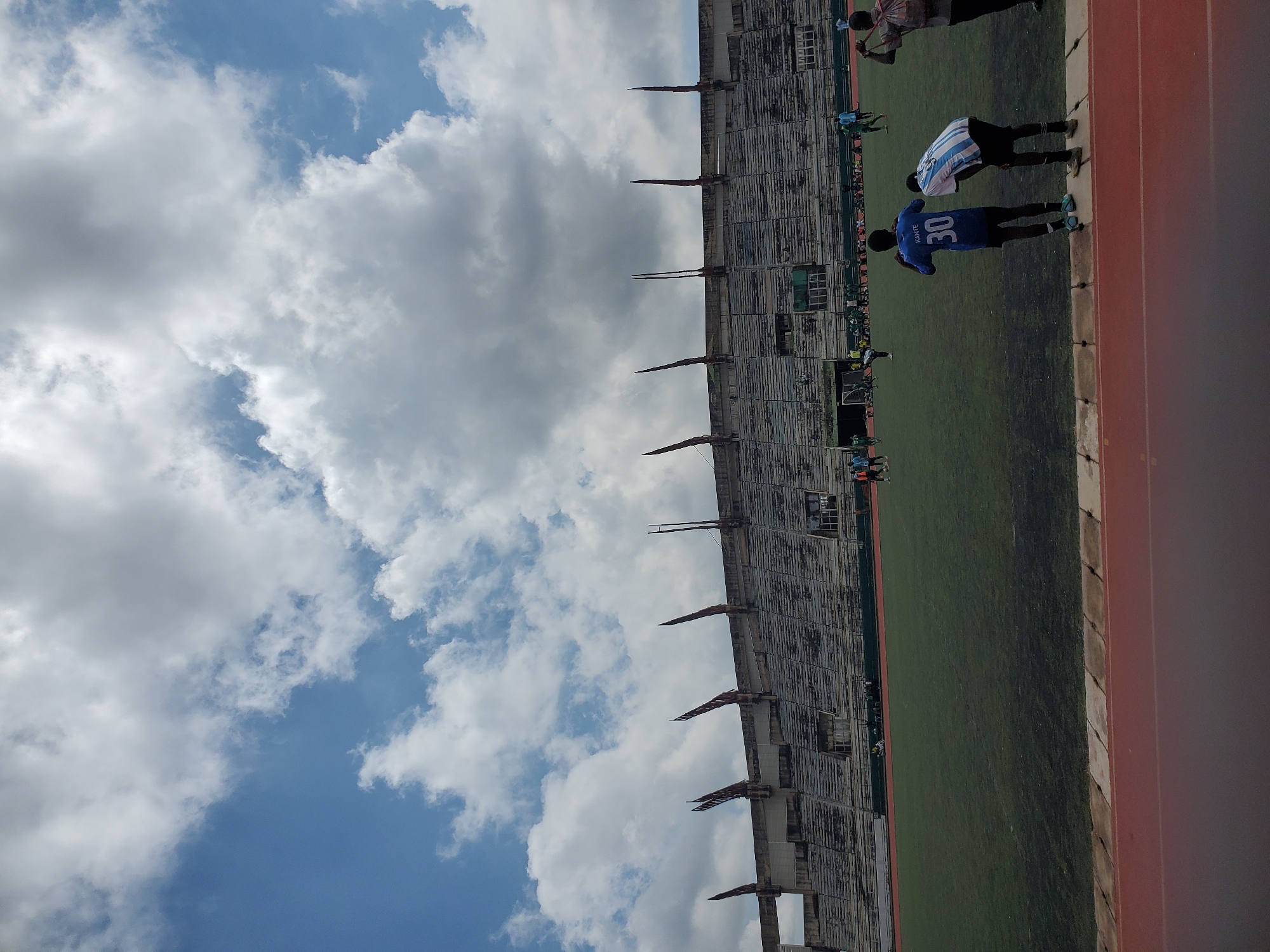The image shows a football stadium in New Stadium, Nigeria, under a partly cloudy sky. In the foreground, two young males in football attire are standing, looking out at the field. One is wearing a blue jersey with the number 30 and "KANTE" across the back, and the other a white and blue striped jersey with the number 5. A third individual, a male, is visible on the far right, facing away from the camera and holding a strap over his shoulder. The field is green with some worn patches, and several other individuals, appearing to be players and possibly spectators, are scattered across it. In the background, the stadium's tiered seating is visible, with a weathered, concrete facade and distinctive pointed structures along the roofline. The overall impression is of an active sports venue on a bright, overcast day.