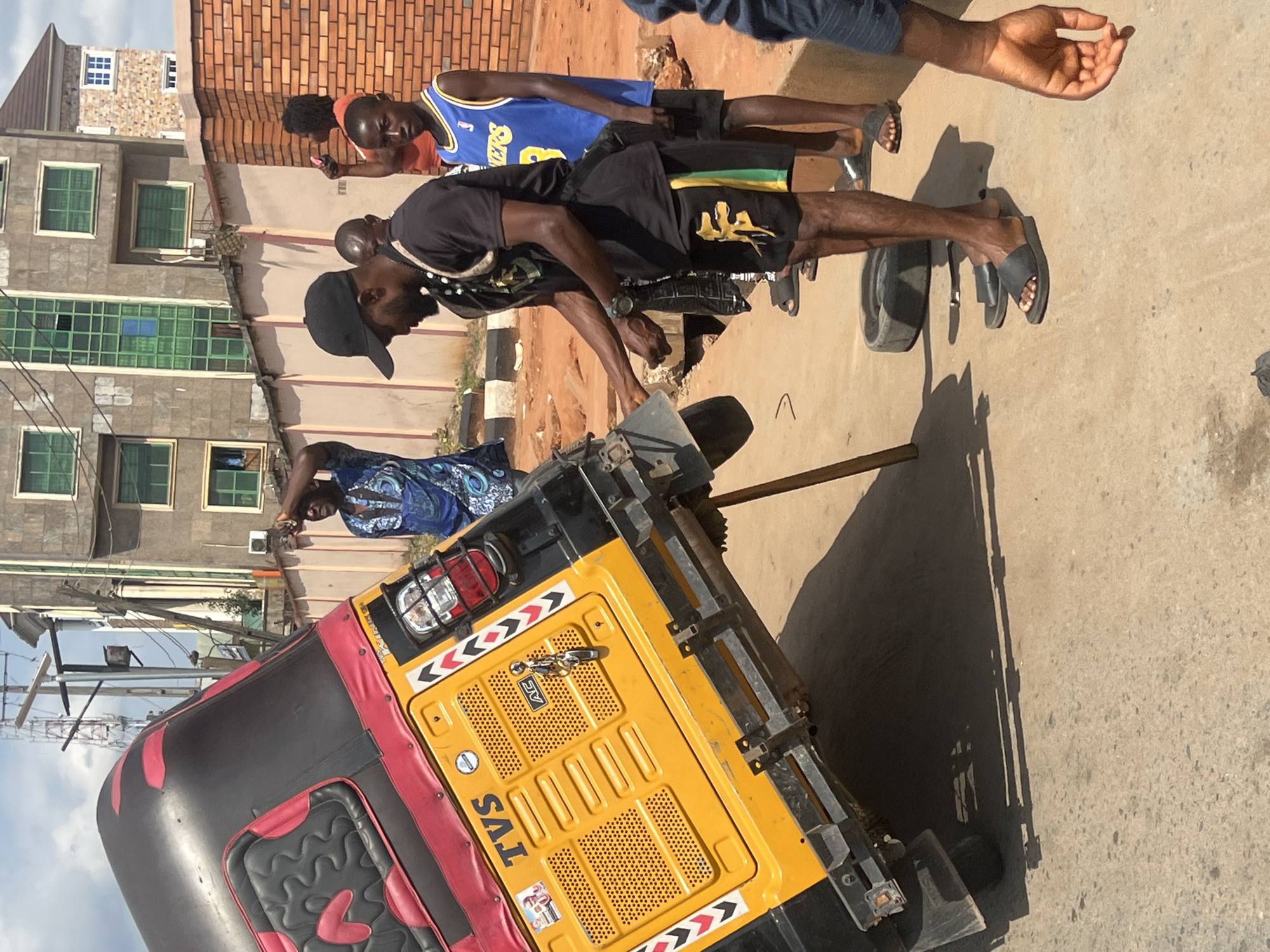 The image depicts a vibrant street scene, likely in an urban or semi-urban setting such as Onitsha, Nigeria. The bright, clear sunlight indicates it is daytime, casting distinct shadows on the concrete ground.
In the foreground, a yellow and black tricycle, commonly known as a keke napep or auto-rickshaw, is prominently featured from its rear. It has red trim around its seating area and visible red accents. The brand name "TVS" is clearly visible on its yellow rear panel, along with a smaller "AG" emblem and black and white chevron patterns near the tail lights. A long, slender pole extends upwards from the rear of the tricycle, suggesting it might be propped up or undergoing some form of inspection or repair.
Several men are gathered around the tricycle. One man, wearing a black t-shirt, black shorts with green and yellow stripes, and a black baseball cap, is bent over, seemingly engaged with the tricycle or something on the ground near its rear wheel. Another man stands nearby, dressed in a blue basketball jersey featuring the "Lakers" logo. Further back, a man in a blue patterned shirt is visible. In the upper right corner, part of a man's arm and hand are seen, reaching into the frame.
The background consists of multiple buildings, some with green-framed windows and others showcasing exposed reddish brickwork, typical of residential or commercial structures in the region. A utility pole is also visible in the distance. The scene captures an everyday moment of activity or a casual gathering on the street.