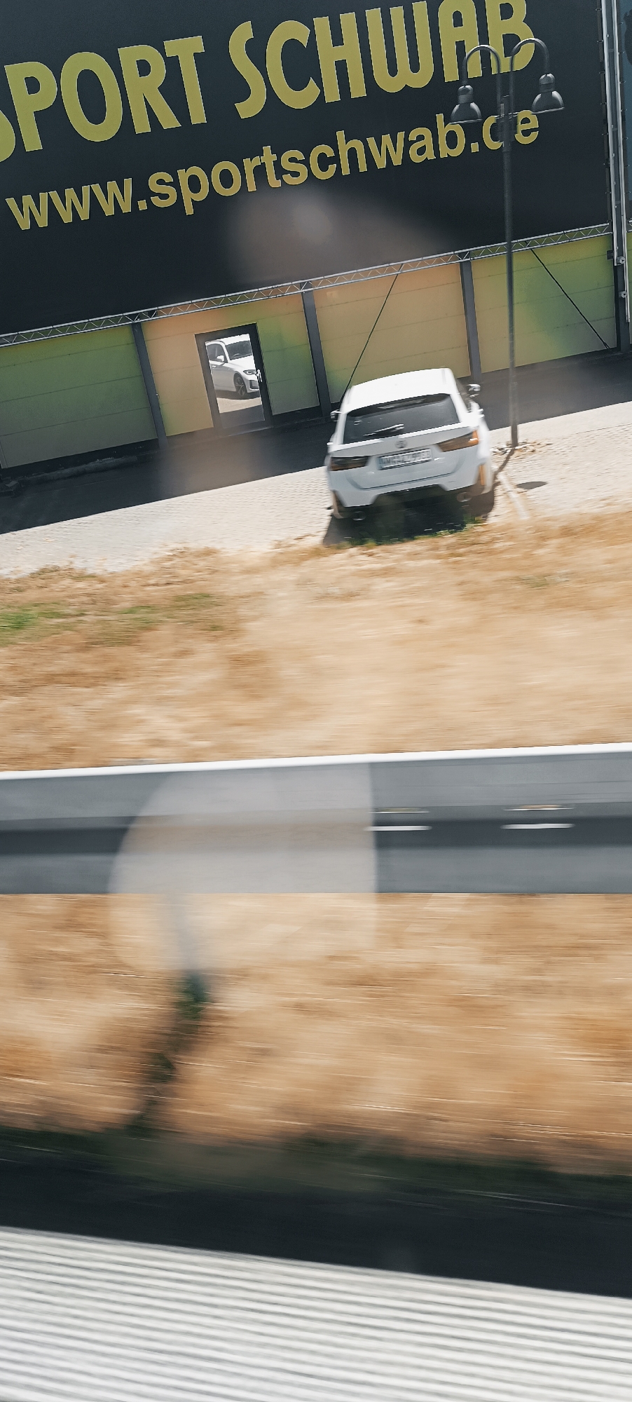 The image depicts a street scene in Winterbach, Germany. In the foreground, a blurry, dry, grassy area is visible, suggesting a roadside or open field. A portion of a modern, grey structure, possibly a building or barrier, runs horizontally across the middle of the frame, with a blurred line marking its edge. Beyond this structure, a paved area is visible, with two white cars. The car on the right is a white station wagon, parked at an angle, showing its rear. A smaller white car is visible through an open doorway of a building in the background, which appears to be a commercial establishment with a dark facade and a yellow and green accent. A large black banner with yellow lettering is prominently displayed above the building, featuring the text "SPORT SCHWAB" and "www.sportschwab.de". A street lamp with two lights is positioned on the right side of the image, casting a shadow. The scene appears to be captured from a moving vehicle, indicated by the motion blur throughout the image.