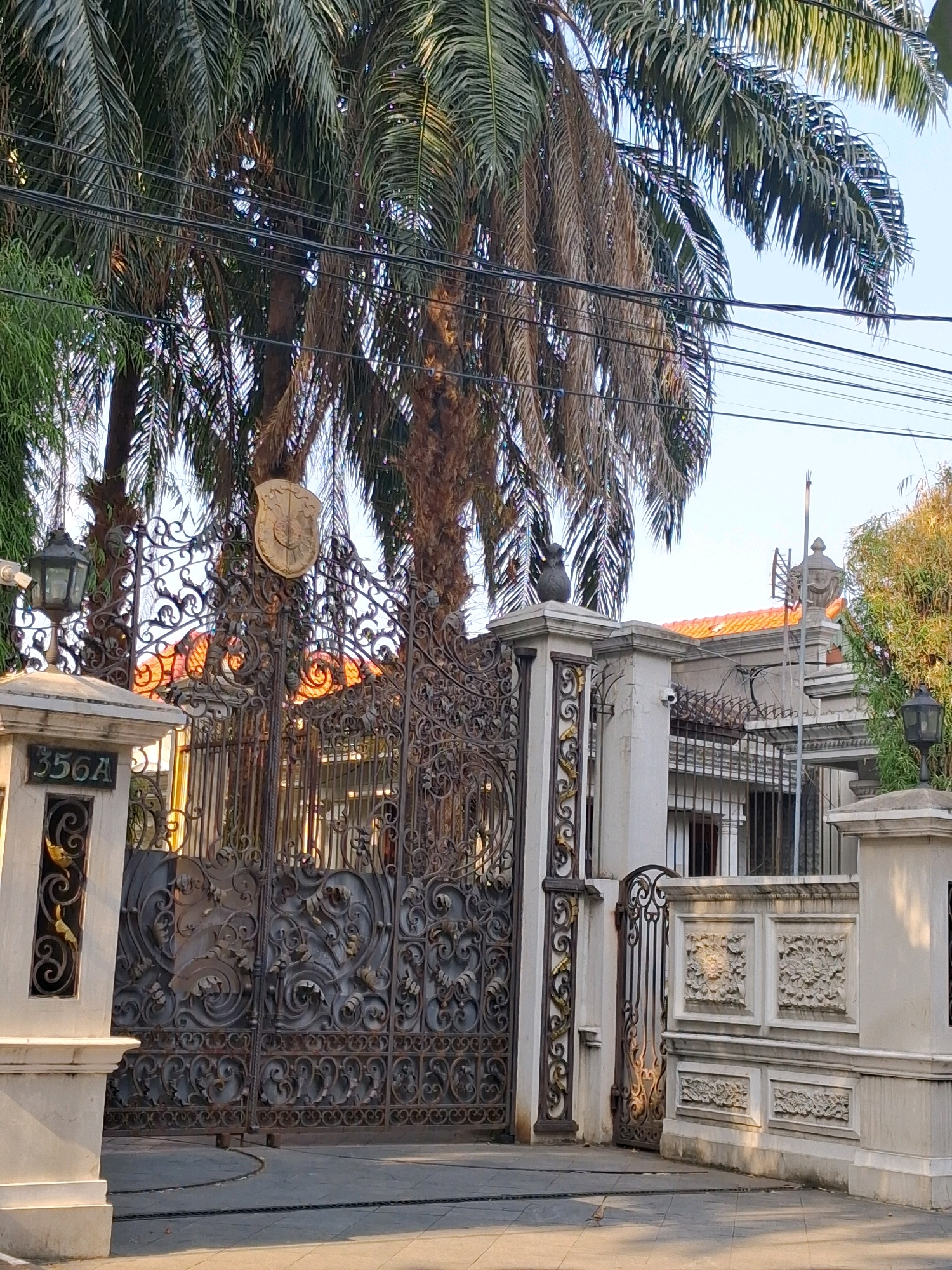 A wide shot captures a grand, ornate wrought iron gate and fence in front of a building in Surakarta, Indonesia. The gate is intricately designed with swirling patterns and floral motifs. Above the gate, a gold crest is mounted. To the left of the gate, a pillar holds a black lantern and a sign that reads "356A". The fence continues to the right, featuring decorative panels with relief carvings of flowers. Behind the fence, the building's facade is visible, with a tiled roof and barred windows. Tall palm trees with fronds partially obscure the sky, which is a clear blue. Power lines stretch across the upper portion of the image. The ground in front of the gate is paved with interlocking stones, and a small bird is visible on the pavement. The scene appears to be captured during daylight.
