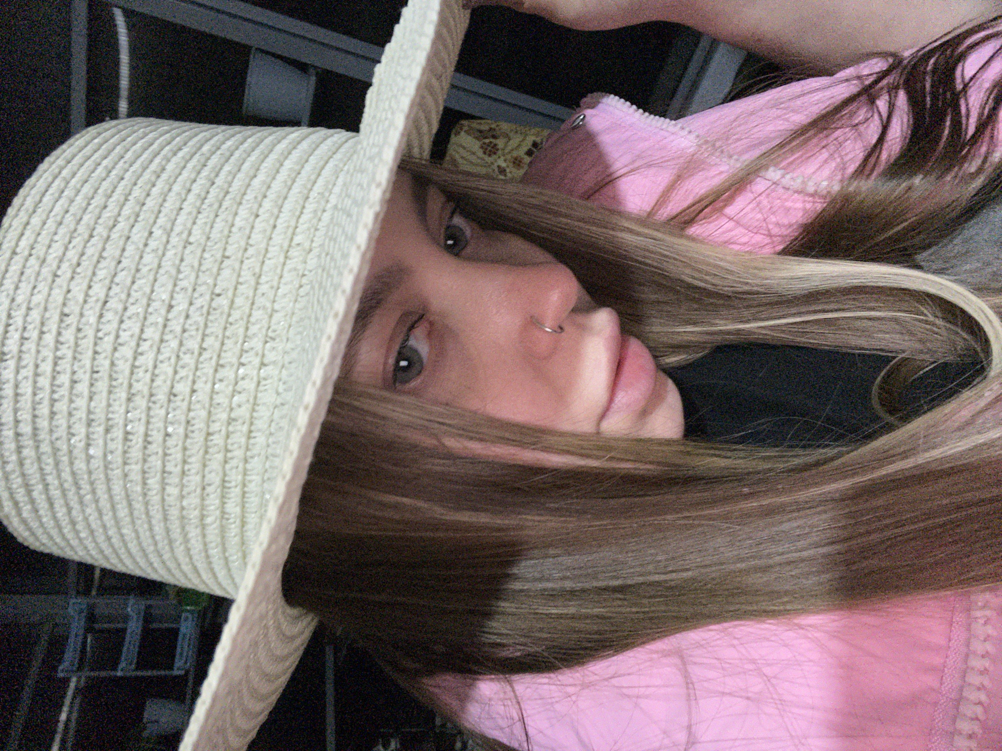 The image is a close-up portrait of a woman wearing a straw hat and a pink jacket. The woman has light brown hair, fair skin, and a nose piercing. The background is dark and indistinct. The image appears to have been taken outdoors, possibly at night or in a poorly lit area. There is no specific information in the image to confirm the location in the city of Novopokrovskaya, Russia.