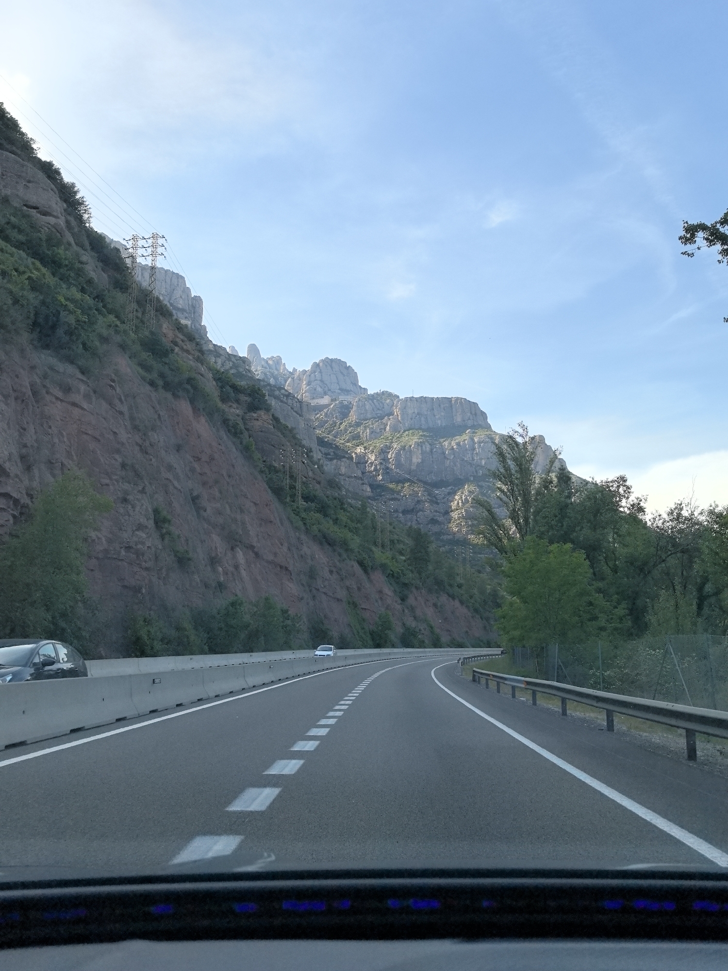 The image presents a view from inside a vehicle driving along a multi-lane highway in a mountainous region, specifically near Collbató, Spain. The road curves gently to the right, featuring white dashed lane markers and a solid white line along the right shoulder. On the left, a rugged and steep mountainside dominates the scene, composed of reddish-brown rock formations interspersed with green foliage. Several electricity pylons and their accompanying power lines traverse the lower slopes of this mountain. Further in the background, the distinctive, jagged peaks of the Montserrat mountain range are clearly visible under a vast, bright blue sky with sparse, wispy clouds. To the right of the highway, a lush green forested area is bordered by a metal guardrail. A sturdy concrete barrier separates the road from the mountain on the left. Three vehicles are captured: a dark car partially visible in the immediate foreground, suggesting the viewpoint from within a car, and two light-colored cars further down the road. The bright, clear sky indicates pleasant weather, likely late morning or early afternoon. The very bottom of the frame shows a portion of the car's dashboard with subtle blue lights. No legible text is visible within the scene.