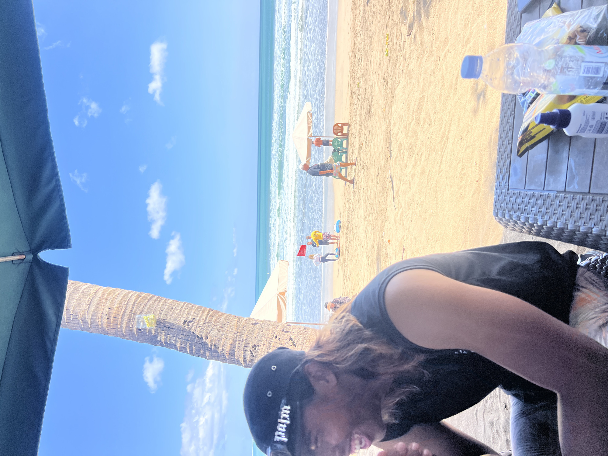 The image shows a beach scene in Kuta, Indonesia, with the Indian Ocean in the background. A man wearing a black t-shirt and a black baseball cap with the word "palm" is in the foreground, laughing. In the middle ground, several people are visible on the sandy beach, some standing under umbrellas and others walking. The ocean is a bright turquoise color with moderate waves. The sky is a clear blue with scattered white clouds. A palm tree is visible on the left side of the image, with a clear plastic bag containing yellow objects attached to it. On the right side of the foreground, a table with a water bottle and a spray bottle is visible. The overall setting appears to be a sunny day at the beach.