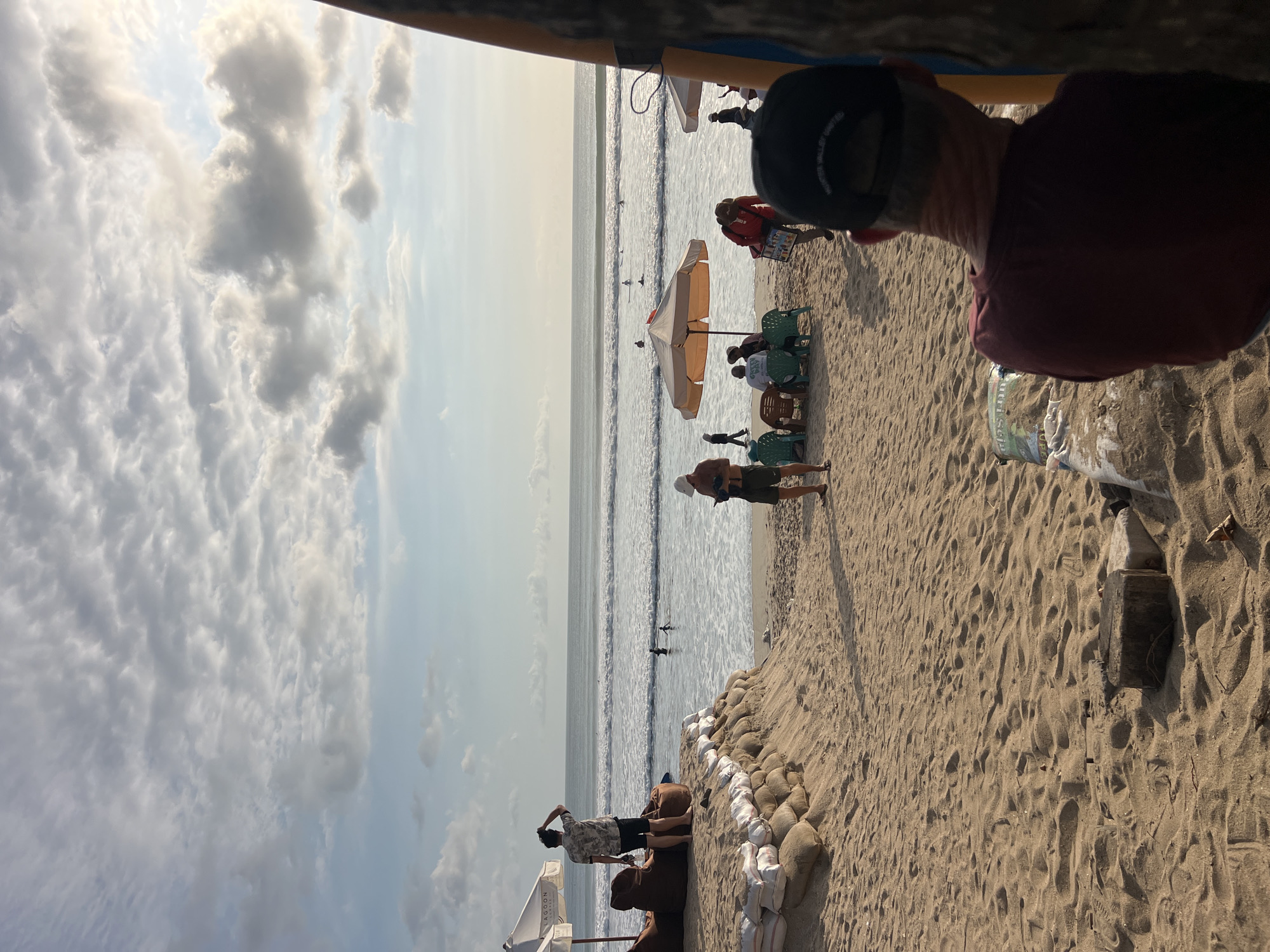 The image depicts a sandy beach scene in Kuta, Indonesia, during what appears to be late afternoon or early evening. The foreground is dominated by textured sand with scattered debris, including a piece of wood and a plastic bottle. In the middle ground, several people are visible. A person in a dark t-shirt and black baseball cap is facing away from the camera, partially obscuring a surfboard on the right. To the left, a sandbag barrier separates the beach from a slightly elevated area where two people are standing near beanbag chairs and umbrellas. Beyond this, the ocean stretches out, with gentle waves and several surfers visible in the water. The sky is partly cloudy, with the sun beginning to set, casting a warm light on the scene. The overall atmosphere is relaxed and tranquil, characteristic of a beach resort area.