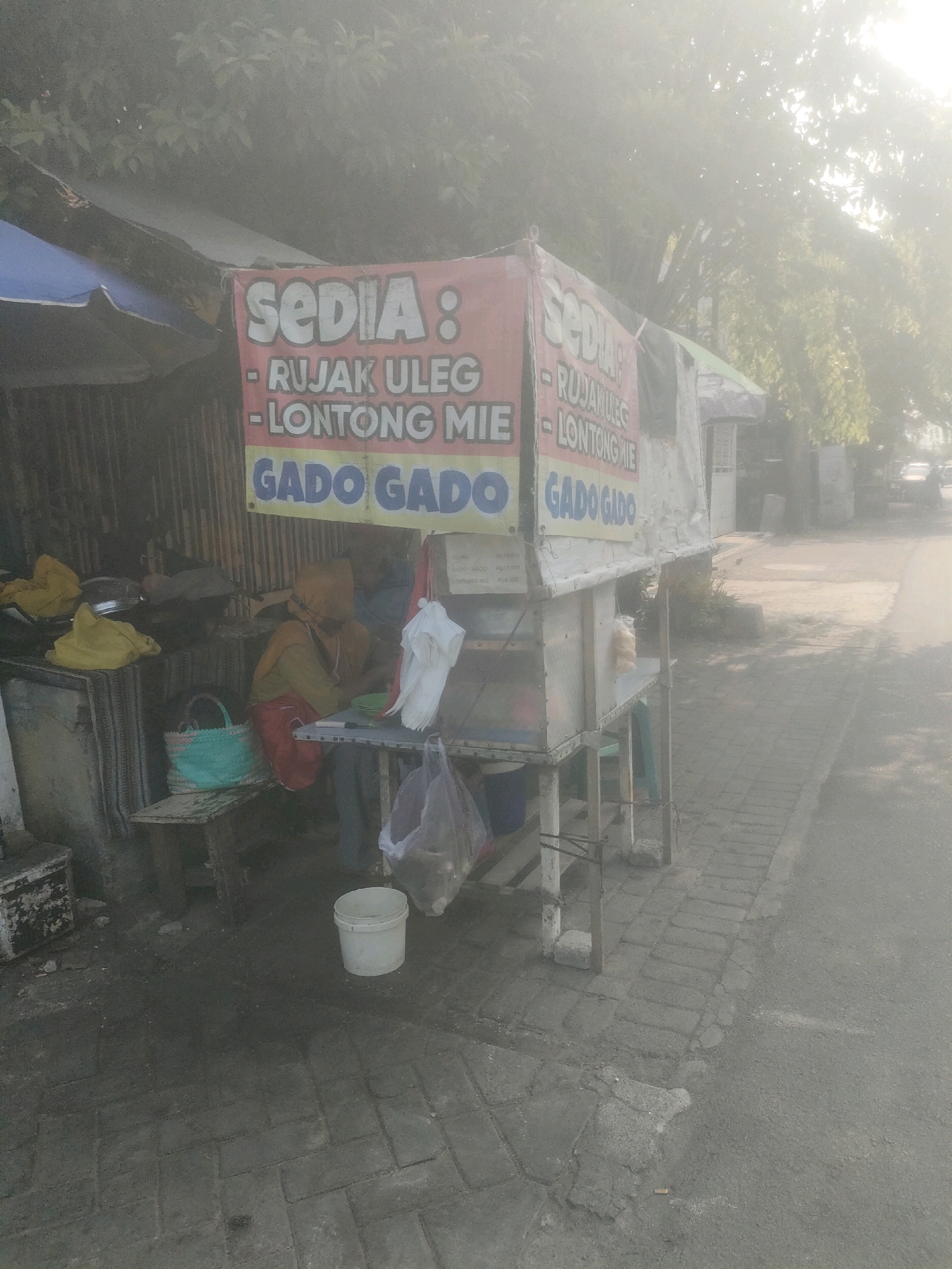 This image depicts a street food stall in Surabaya, Indonesia, during what appears to be a hazy or misty morning. The stall is set up along a paved sidewalk next to a road. The stall itself is a mobile cart with a tarp roof and a banner that reads "SEDIA: RUJAK ULEG, LONTONG MIE, GADO GADO."
A person, likely the vendor, is seated behind the counter, partially obscured by the haze and a headscarf. They appear to be preparing food, with bowls and utensils visible on a small table. Various food items and supplies are stored on shelves and around the stall, including bags of ingredients, a turquoise woven basket, and a white bucket. The background shows trees and foliage, with some buildings and vehicles faintly visible further down the road, indicating an urban or suburban setting. The overall atmosphere is diffused due to the haze, softening the colors and reducing the clarity of distant objects.