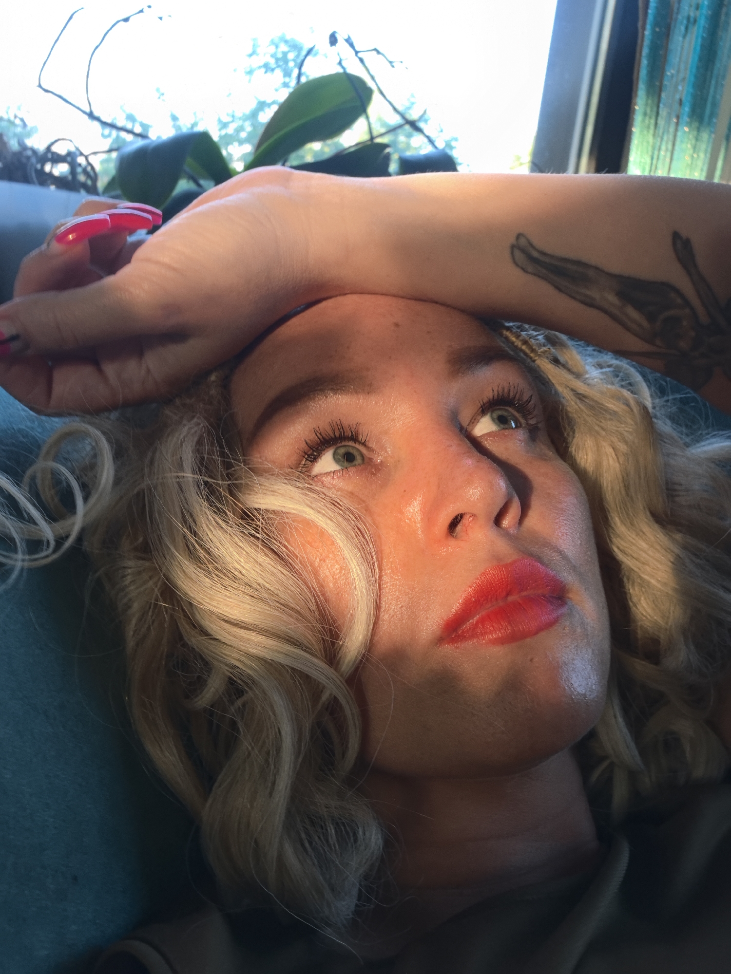 The image is a close-up, low-angle shot of a woman lying down, with her face turned upwards and to the right, looking out of frame. Her left arm is bent and resting on her forehead. She has blonde, curly hair, bright green eyes, and is wearing bright red lipstick. A tattoo is visible on her forearm, and her fingernails are painted pink. Sunlight streams in from the right, casting shadows across her face. In the background, there are green leaves from a plant and a window with a beaded curtain. The scene appears to be indoors, possibly in a residential setting, with no specific identifiable landmarks of Noviy, Russia.
