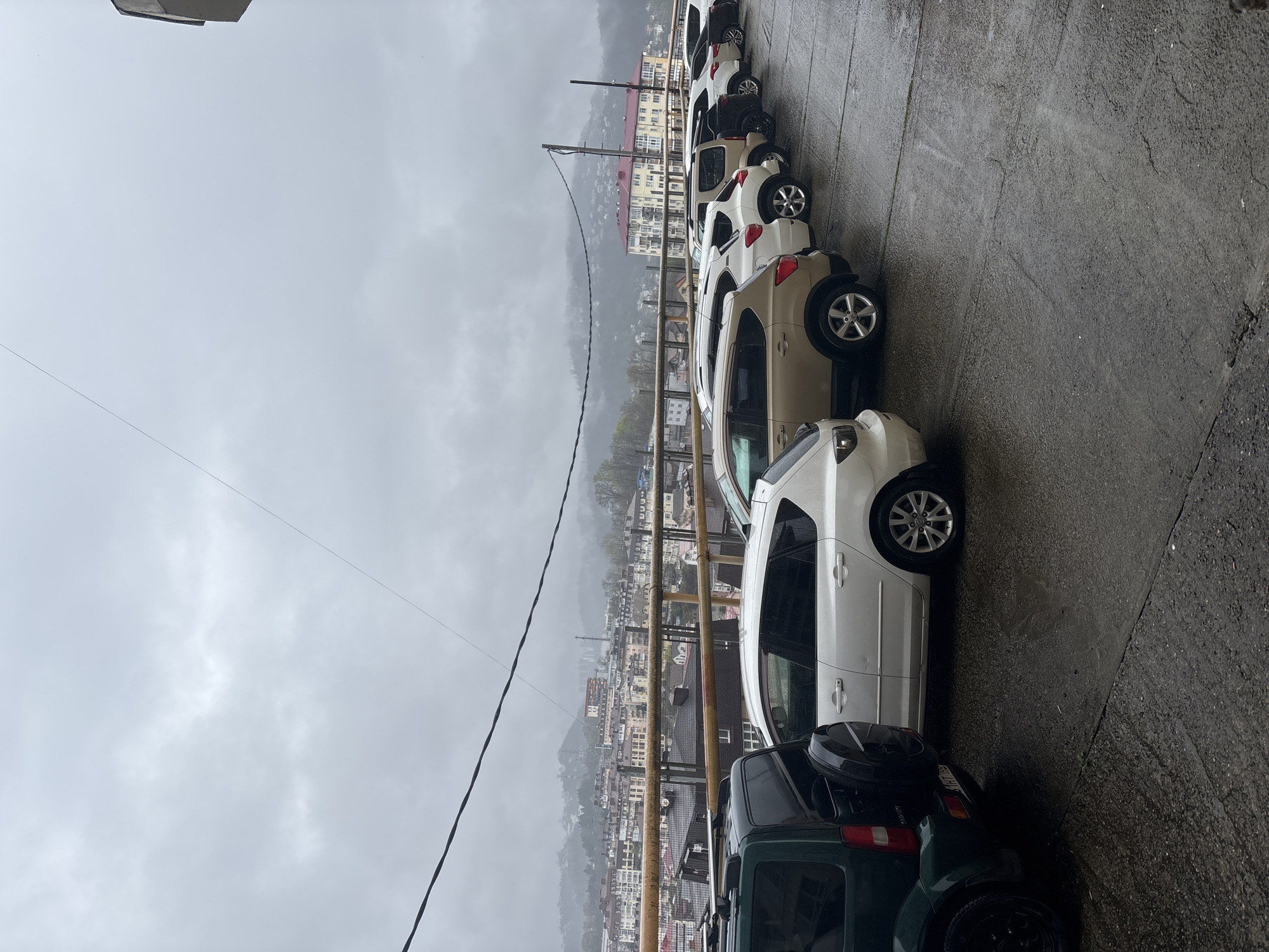 The image shows a street in Sochi, Russia on an overcast day. A row of parked cars lines the wet asphalt road, angled uphill. The vehicles are various makes and colors, including dark green, white, and tan. In the background, a cityscape extends towards the mountains, densely packed with buildings. Utility poles with power lines run along the side of the street.