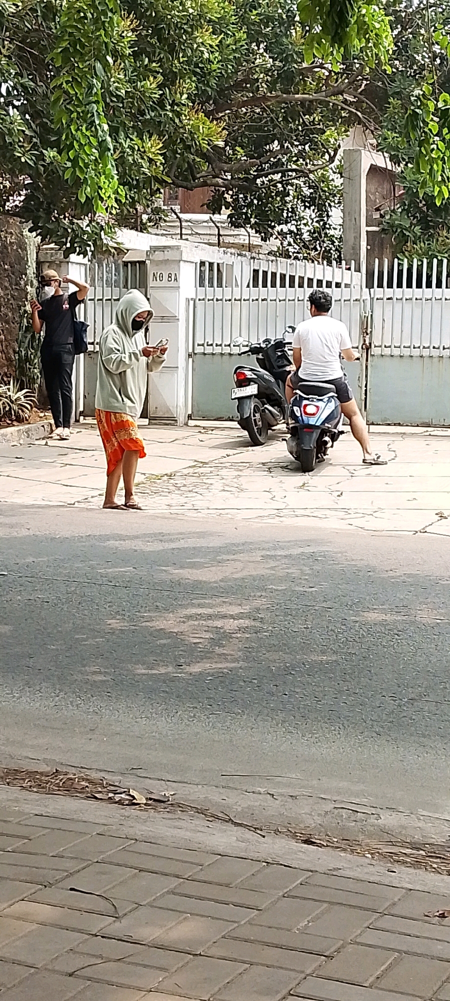 In Depok, Indonesia, on a sunny day, three individuals are present on a paved street. The street is bordered by a sidewalk and a gate of a building with the inscription "NO 8A". A person in a light gray hooded sweatshirt and orange patterned shorts, wearing a black face mask, stands in the middle of the scene, looking down at a mobile phone held in their hands. To the left, another person, wearing a dark t-shirt, black pants, a beige cap, and a white face mask, stands near a wall, with their right arm raised as if gesturing or adjusting their cap. To the right, a person is seated on a light blue and black scooter, facing away from the camera, wearing a white t-shirt and dark shorts. Another dark scooter is parked behind the first. Green foliage from trees overhangs the scene from the top. The ground is a mix of cracked asphalt and interlocking paving stones.