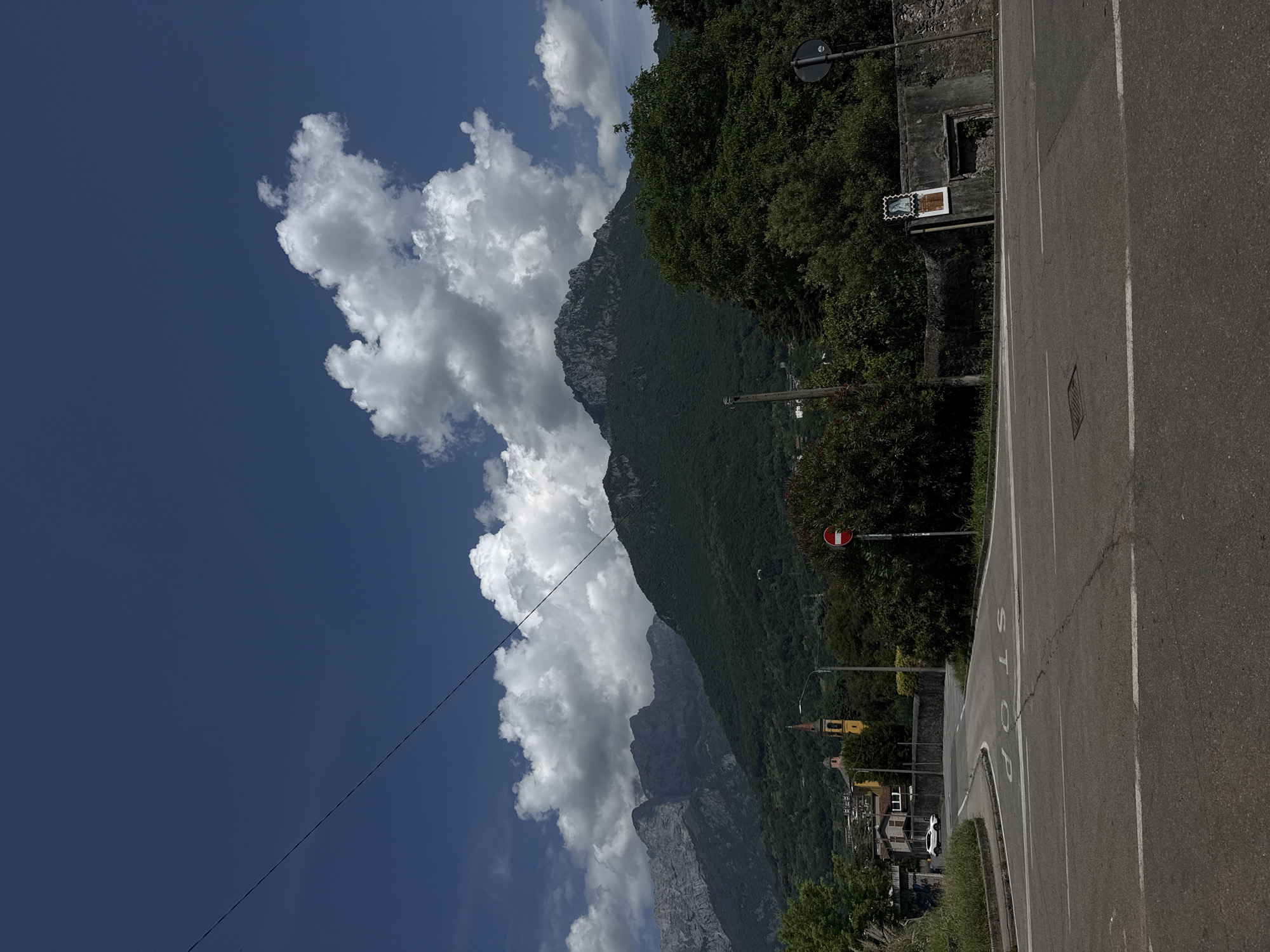 The image depicts a rural or suburban landscape under a clear blue sky with scattered white clouds. A prominent feature is a large, dark green mountain covered in trees, with a more rugged, rocky peak visible in the distance. In the foreground, there is an asphalt road with white lane markings and a visible "STOP" sign painted on the road surface. To the left of the road, there is a white car parked near a cluster of buildings, one of which has a yellow bell tower and a red roof. A power line runs diagonally across the upper portion of the image. Dense green foliage and trees line the right side of the road, and a low stone wall separates the road from the vegetation. A circular red and white traffic sign is visible on the right side of the road.