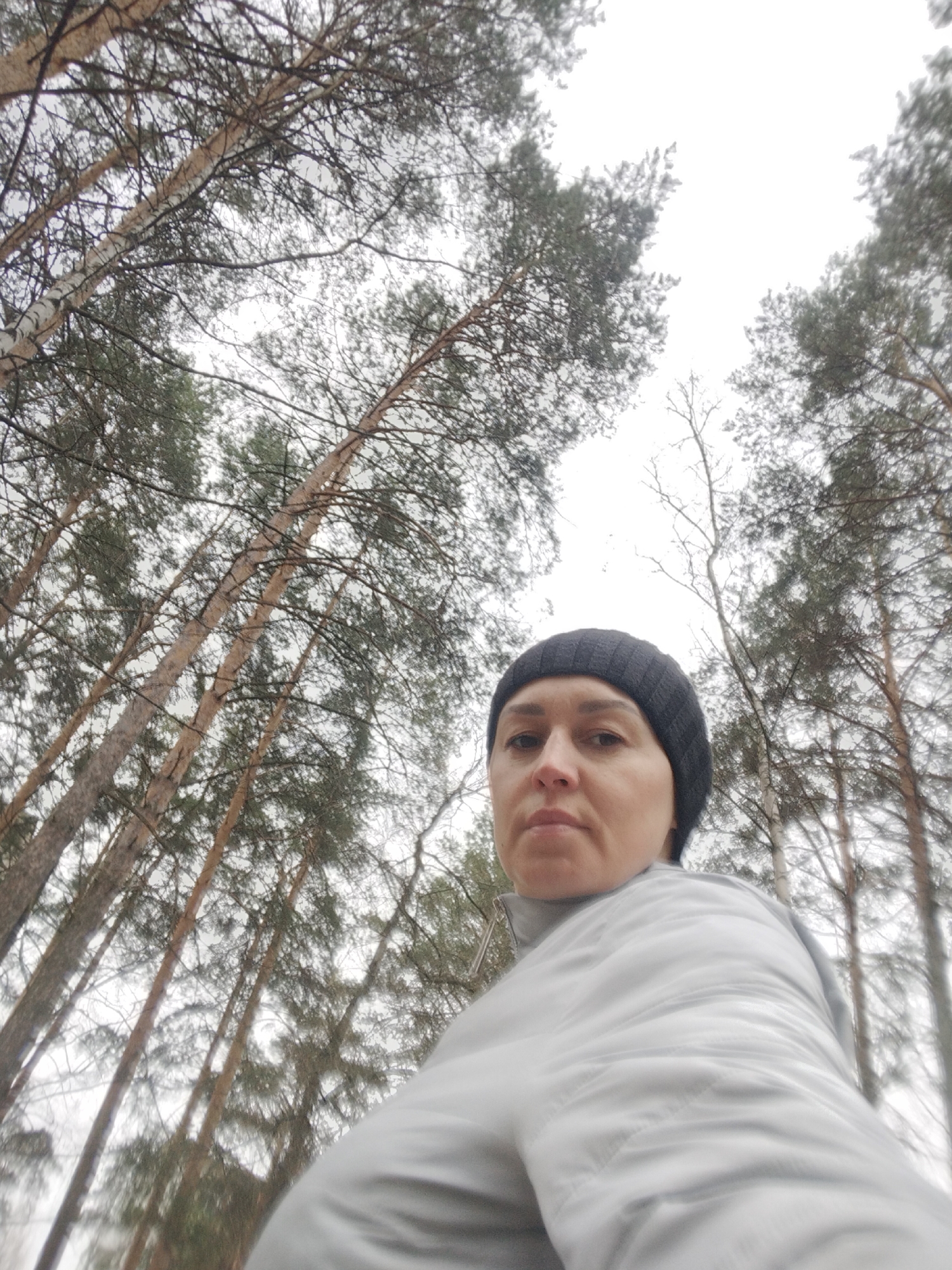 A low-angle outdoor portrait features a woman standing in a dense forest, looking directly at the camera. She wears a dark ribbed beanie and a light grey or off-white jacket, indicating cool weather. Her expression is neutral.
The background is filled with numerous tall trees, stretching upwards towards an overcast sky. Prominently visible are both birch trees, identifiable by their light, often white, bark and delicate bare branches, and coniferous trees, likely pines, with their darker, needle-like foliage. The dense arrangement of trees and their upward angle creates an immersive forest atmosphere. The sky appears bright but cloudy, diffusing the light. The scene depicts a natural, wooded environment, likely during the daytime. This setting is located in Chaikovsky, Russia. No visible text is present in the image.