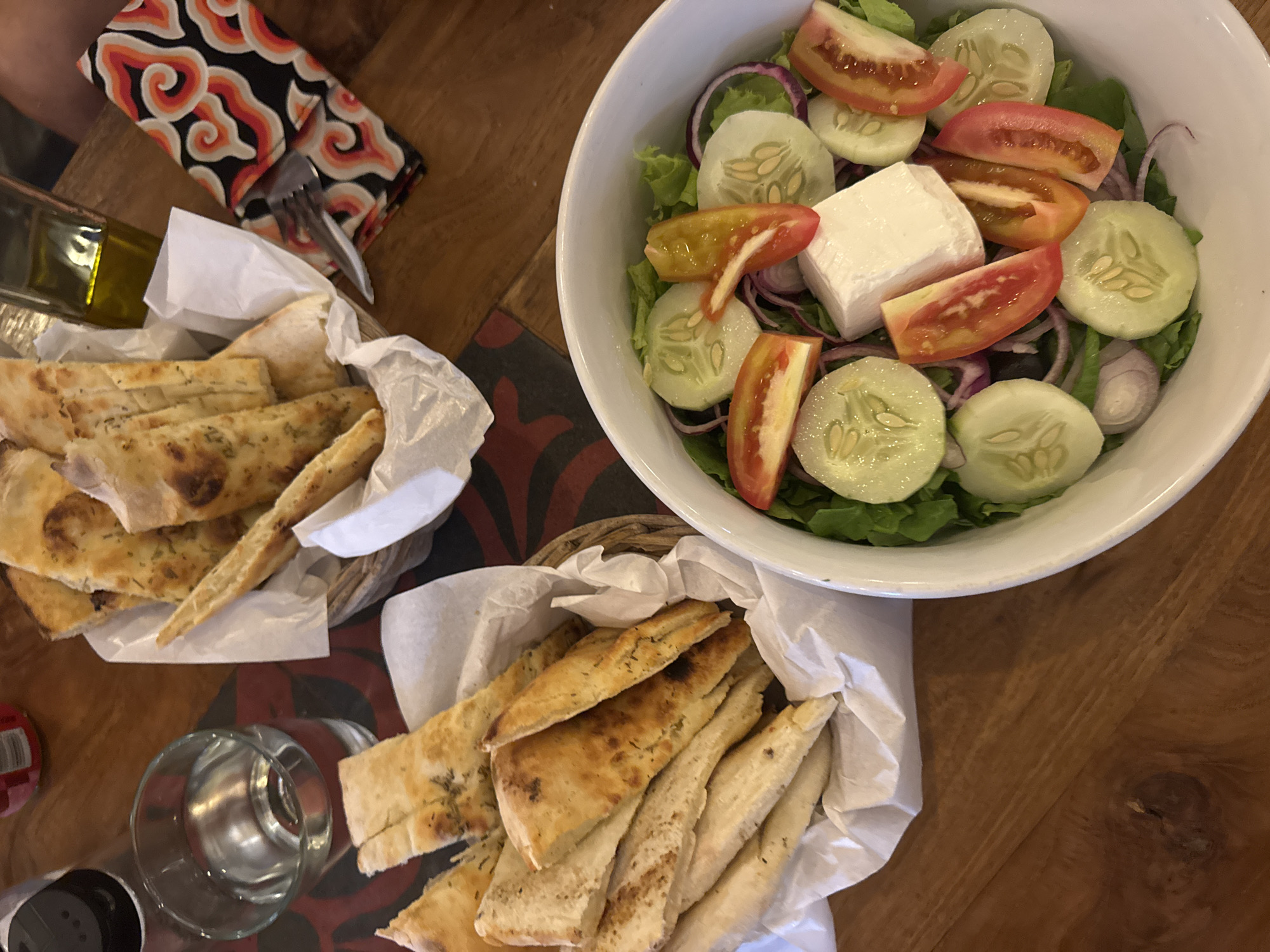 A still life on a wooden table depicts a meal, seemingly at a restaurant in an unknown city. On the right side, there is a large white bowl containing a salad consisting of lettuce, cucumber slices, tomato wedges, red onion rings, and a block of white cheese, likely feta. To the left are two wicker baskets lined with white paper. Each contains slices of flatbread, possibly focaccia, with visible herb seasonings. A folded napkin with a colorful swirling design and cutlery are partially visible near one basket, along with a small bottle of olive oil. A drinking glass and a small can are located on the left side near the bottom basket. The scene suggests a casual dining experience.