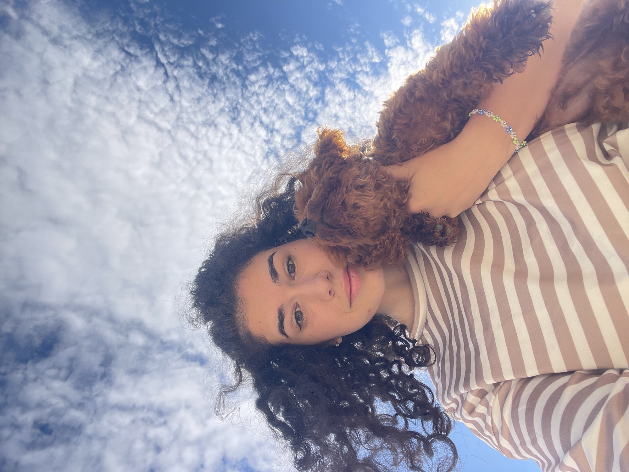 The image displays a young woman holding a small, brown, curly-haired dog outdoors in Koprinka, Bulgaria, against a backdrop of a bright blue sky interspersed with white, scattered clouds. The woman, positioned in the lower center of the frame, has dark, curly hair, brown eyes, and a neutral expression with a slight smile. She is wearing a long-sleeved shirt with horizontal stripes in alternating shades of light brown/tan and white. On her right wrist, a delicate beaded bracelet composed of small light blue, light green, and white beads is visible. The dog, appearing to be a poodle or doodle breed, is held close to the woman's upper chest and shoulder, with its head resting near her right shoulder and its eyes appearing closed or directed upwards. The image is captured from a low-angle perspective, looking upwards at the subjects with the sky dominating the upper two-thirds of the composition.