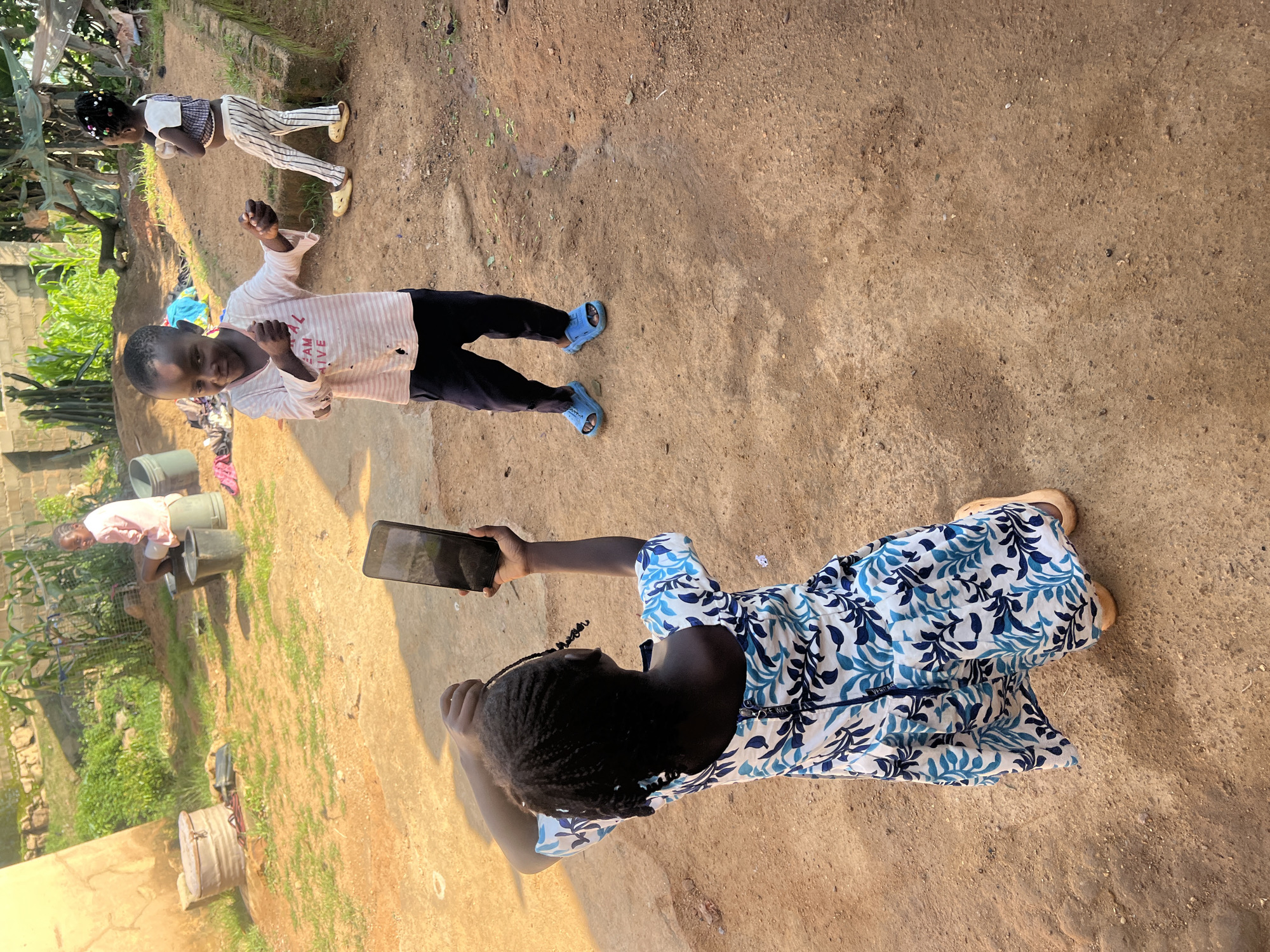 In the city of Jos, Nigeria, three children are visible in an outdoor, dusty, dirt-yard setting. The child in the foreground, facing away from the camera, wears a blue and white floral dress and holds a black smartphone up to take a selfie. To their left, a second child, facing the camera and smiling, wears a pink and white striped long-sleeved shirt and dark blue pants, with blue sandals on their feet. To the right and slightly behind, a third child, wearing a black and white striped outfit and yellow sandals, stands with their back to the camera. In the background, an adult male in a pink shirt is seated near buckets and various items, including a large barrel. The environment appears rural, with vegetation and some structures visible in the distance.