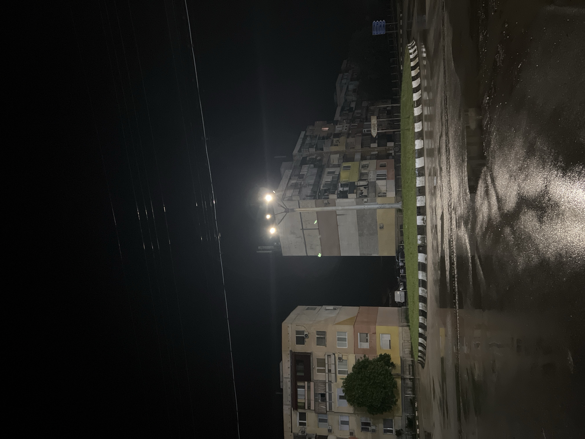 The image depicts a nighttime scene in what appears to be an urban environment in Roman, Bulgaria. The foreground consists of a wet, reflective road surface. A black and white striped curb separates the road from a narrow strip of grass and nearby buildings. These structures appear to be residential buildings, possibly apartments. The buildings exhibit a mix of colors and architectural styles. Streetlights illuminate the area, casting a glow on the buildings and the wet pavement. Power lines are visible against the dark sky. The overall scene suggests a quiet, rain-soaked evening in a residential part of Roman.