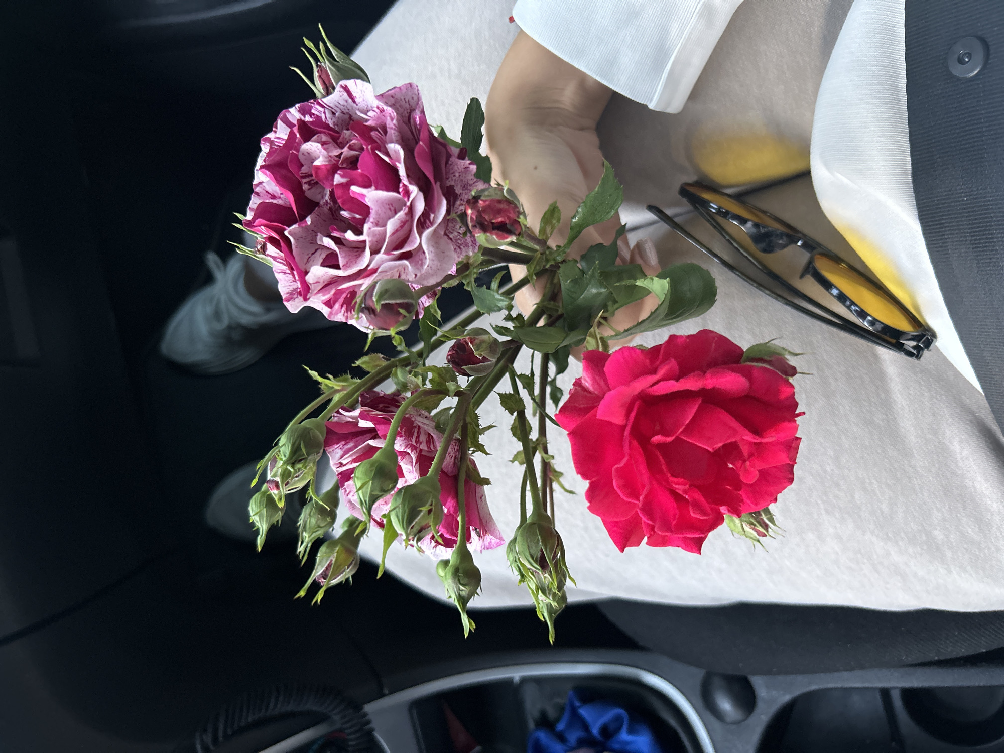 The image shows a close-up view of a hand holding a bouquet of roses, likely inside a car. The bouquet features two prominent roses: one a deep red and the other a mix of pink and white with a speckled pattern. There are also several smaller buds in various stages of development attached to the stems. The hand holding the flowers is fair-skinned, and a portion of a white garment is visible behind it. Adjacent to the flowers are a pair of sunglasses with dark frames and yellow-tinted lenses. The background of the image shows the interior of a car, including a storage compartment and a glimpse of a passenger seat with a shoe visible. The location is unspecified.