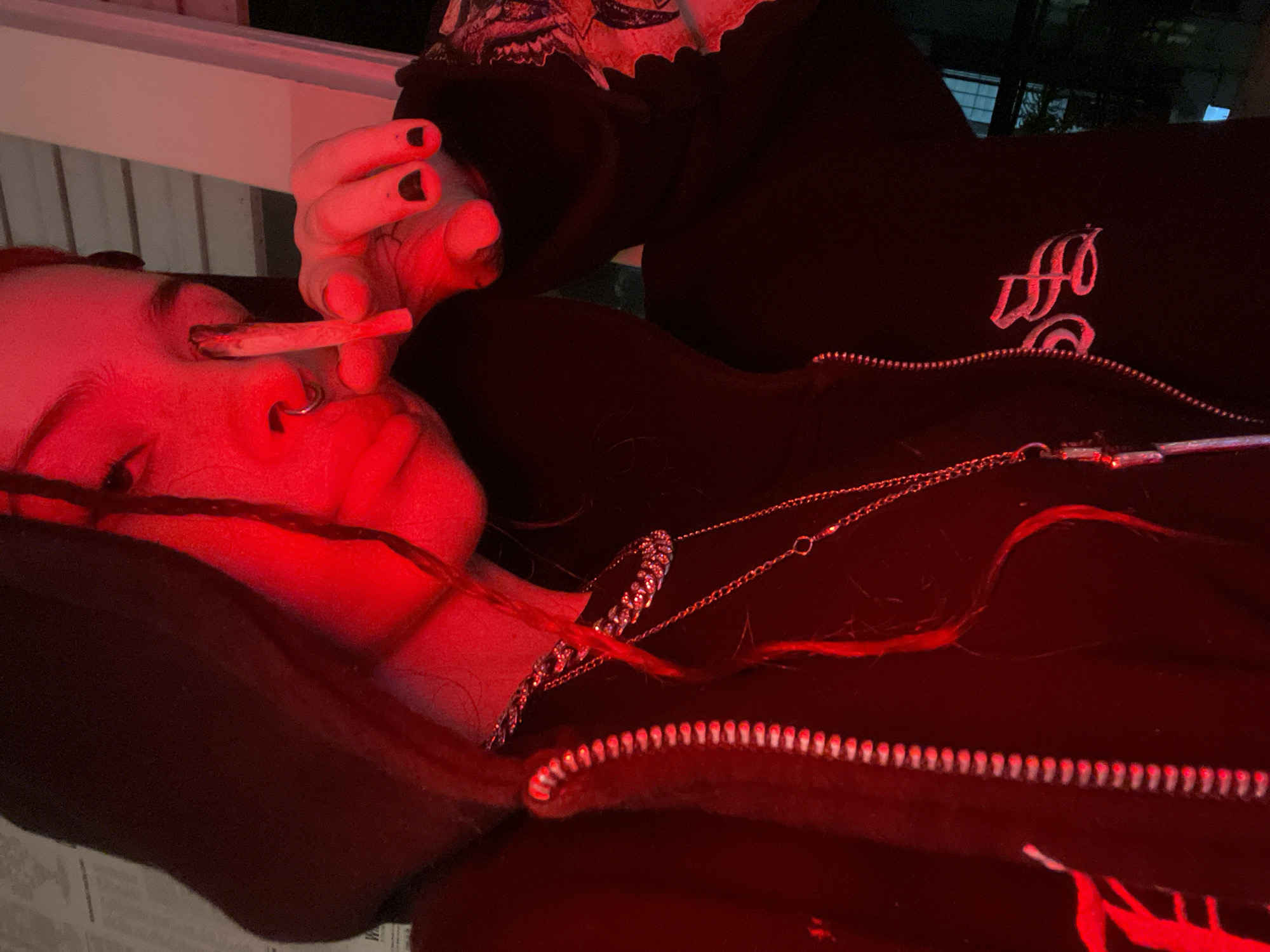A person is holding a lit cigarette close to another person's face under red lighting. The person closest to the camera has a nose ring, and both are wearing dark clothing. The setting appears to be indoors, possibly at night. There is no information to suggest the location is in Buenos Aires, Argentina.