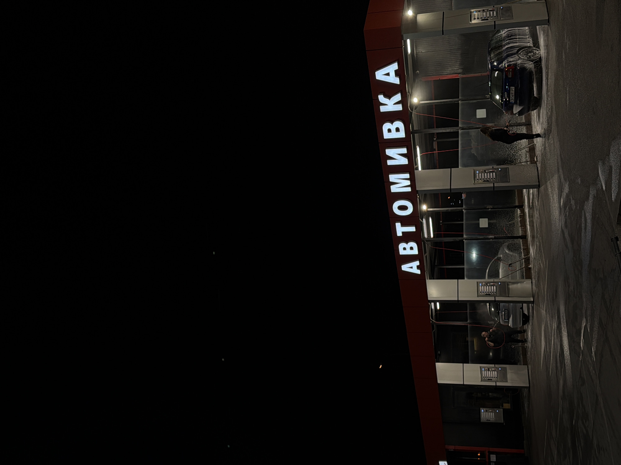 The image captures a nighttime scene at a self-service car wash in Sofia, Bulgaria. The car wash facility features a modern design with individual bays, illuminated by bright overhead lights. The primary structure is clad in a dark red material, displaying prominent white, illuminated Cyrillic letters that spell out "АВТОМИВКА" (AVTOMIVKA), which translates to "car wash."
Two individuals are actively engaged in washing their vehicles within separate bays. In the bay to the right, a person dressed in dark clothing is operating a high-pressure spray wand, directing a stream of water and white foam onto a dark-colored car. This car is heavily covered in soap, indicating an ongoing wash cycle. Its license plate, "CB 5631 PT", is partially visible, with "CB" being a common identifier for Sofia. Another individual is seen in the adjacent bay on the left, also holding a spray wand, suggesting they are washing another vehicle, though that car is less distinctly visible.
The ground surrounding the bays is wet and reflective, indicating the active use of water and soap, and there are visible streaks of water and foam. The overall setting is brightly lit against a dark night sky, highlighting the car wash operation. The scene portrays a practical, everyday activity occurring after dark.
