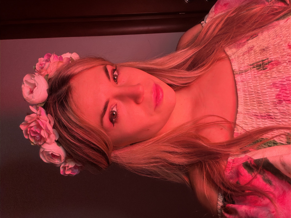 A frontal portrait captures a female individual with blonde hair, illuminated by prominent red light. She wears a floral crown featuring multiple light pink and white artificial peonies. Her long hair is styled down, partially covering her shoulders. Her attire consists of an off-the-shoulder top with a smocked bodice and a floral pattern on the sleeves. Her face exhibits light-colored eyes, visible eyeliner, and bright pink lipstick, with a direct gaze towards the camera. The background is dark and indistinct, characteristic of an indoor environment, with a dark vertical line visible on the right side of the frame, possibly indicating a doorframe or wall edge. The image provides no specific visual information to determine a location within the city of Budva, Montenegro.