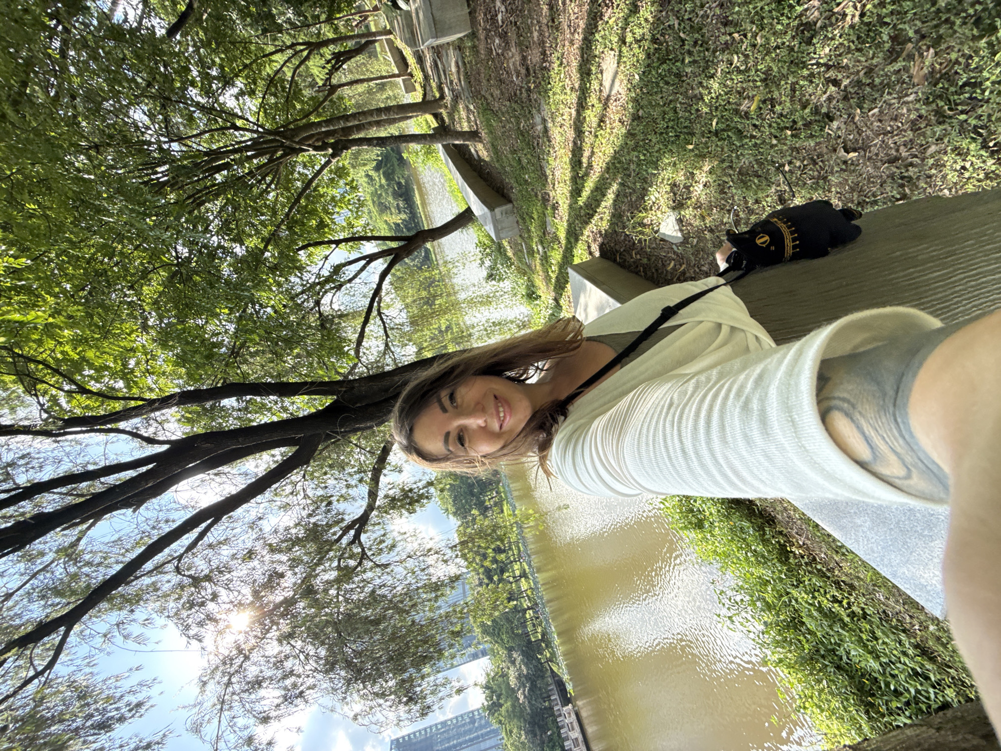 A selfie is taken from a woman's perspective in an outdoor park setting. The woman is smiling and looking at the camera. She is wearing a white cardigan over a green top and has a black bag with a cat design hanging from her shoulder. Her left arm, with a tattoo, is visible in the foreground.
The background shows a body of water with ripples, lined with lush green vegetation and trees. In the distance, several tall buildings are visible, indicating an urban environment. The sun is shining, casting shadows on the grassy ground, and the sky is partly cloudy. Concrete benches are placed along the edge of the water. The location is identified as a park in the city of Huizhou, China.