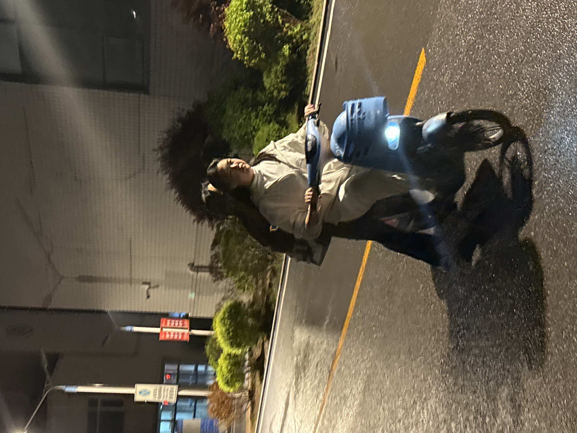 A person is sitting on a blue electric scooter parked on an asphalt road in Zhongtun, China. The road has a yellow dividing line. Buildings and greenery line the side of the road. It appears to be nighttime. The road surface is wet.