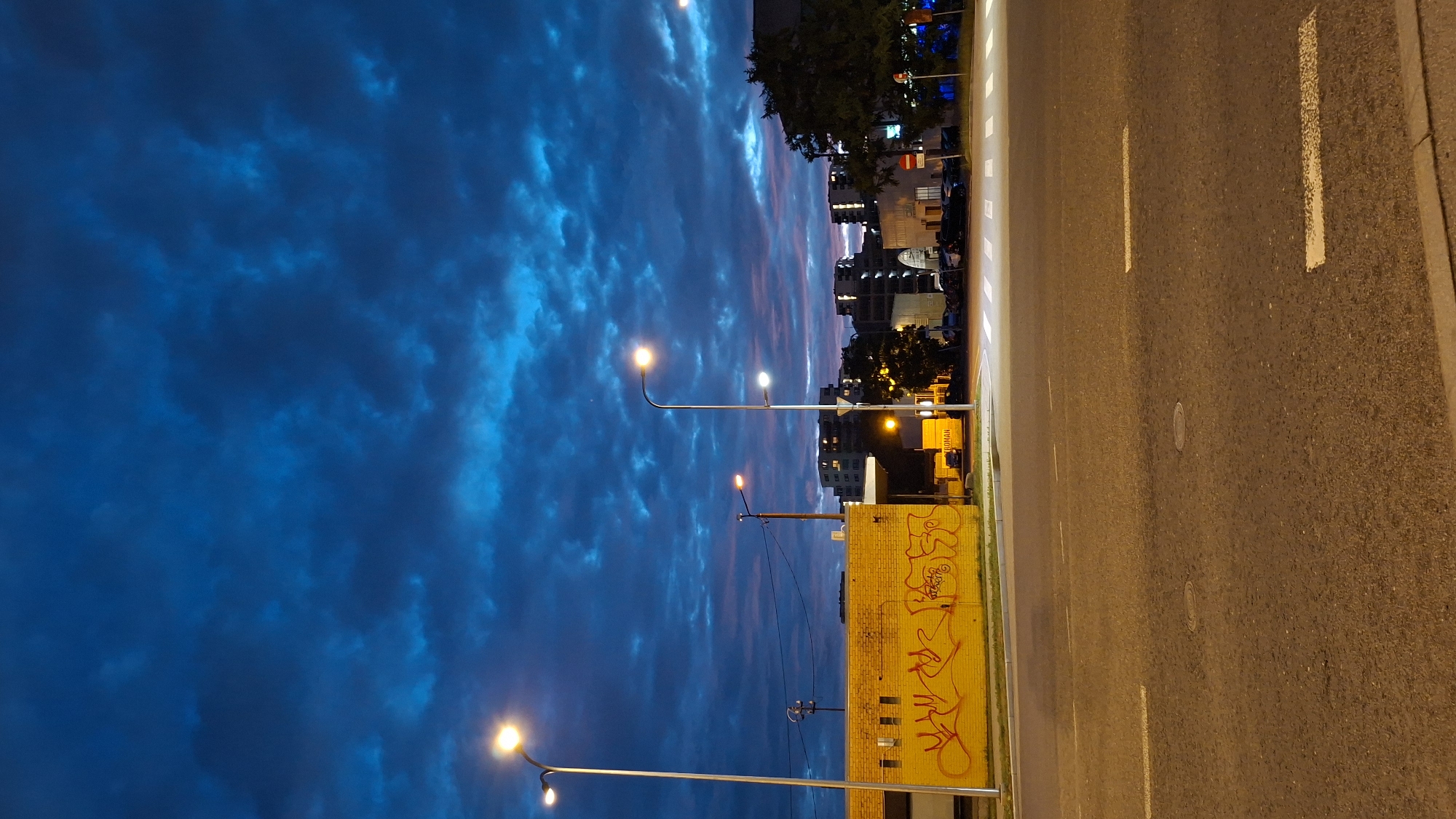 The image depicts a twilight scene in Tallinn, Estonia, focusing on a street in front of a yellow brick building covered in red graffiti. Two streetlights illuminate the area, one on the left and another on the right, with a third, smaller streetlight further in the background. The sky is a dramatic deep blue with textured, low-lying clouds tinged with pink and purple, indicative of sunset or dawn. Beyond the graffiti-covered building, several multi-story apartment buildings and commercial structures are visible, some with illuminated windows. Cars are parked along the street and in front of the buildings, and a pedestrian crossing is marked on the asphalt road in the foreground. The overall impression is an urban environment transitioning from day to night, with an emphasis on atmospheric lighting and architectural elements.