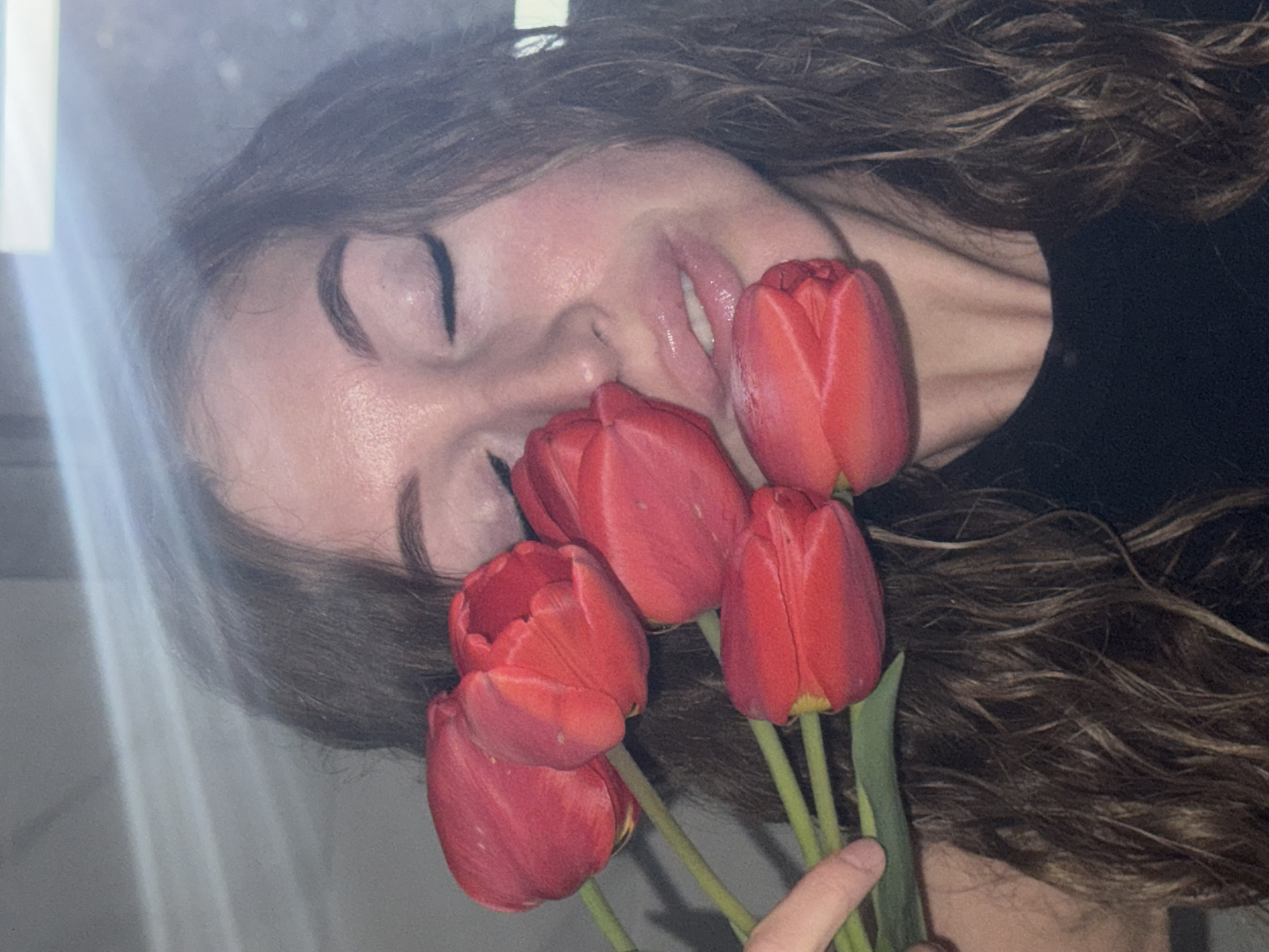 A young woman with brown wavy hair is shown holding a bouquet of red tulips near her face. Her eyes are closed and her lips are slightly parted. She appears to be wearing a black shirt. The background is out of focus and does not provide enough detail to ascertain the location as Chaikovsky, Russia or any other specific place.