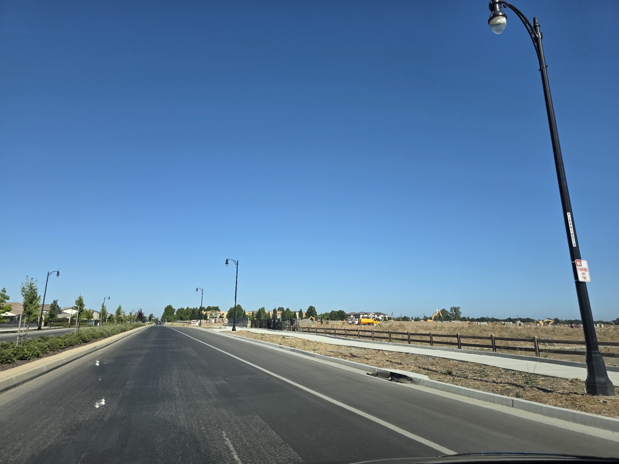 This media file captures a scene on a sunny day in Elk Grove, United States of America. The image shows a paved road with a white line marking the lane. To the left of the road, there is a sidewalk and landscaping with trees and bushes. Across the road, a wooden fence separates the road from a large, undeveloped field. In the distance, there are signs of construction activity, including yellow construction vehicles and several buildings under construction. The sky is a clear, vibrant blue, suggesting pleasant weather. There are several lampposts lining the road, typical of a suburban or developing area. One lamppost in the foreground has a "NO PARKING ANY TIME" sign attached to it. The overall impression is of a developing neighborhood on a bright, clear day.