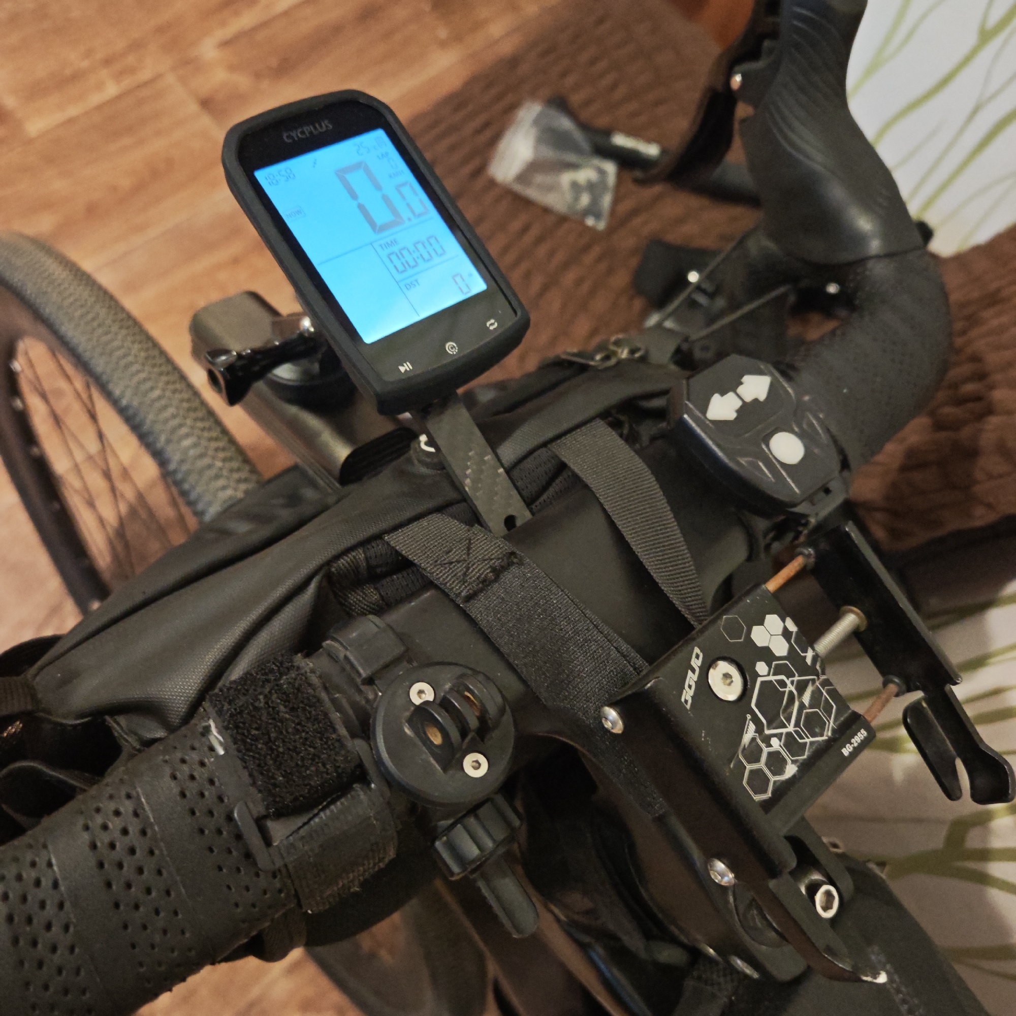 Here is a detailed description of the image:
**Subjects and Objects:** The primary focus is on the handlebars and front part of a bicycle. A CYCPLUS bike computer is mounted on the handlebars, displaying "0.0" for speed and "00:00" for time. The bicycle is equipped with a black handlebar bag secured with straps. There's a device with directional arrows on the handlebars, and a component labeled "GGUO BG-2955" is also visible. A small bag with components is in the upper part of the image.
**Environment:** The background is blurred and includes a hardwood floor and what appears to be a couch or bed with patterned fabric.
**Actions/Interactions:** There is no indication of a person present in the frame, only equipment.
**Location:** There is no visible location in the picture. There is nothing that indicates the city of Rostov in Russia.