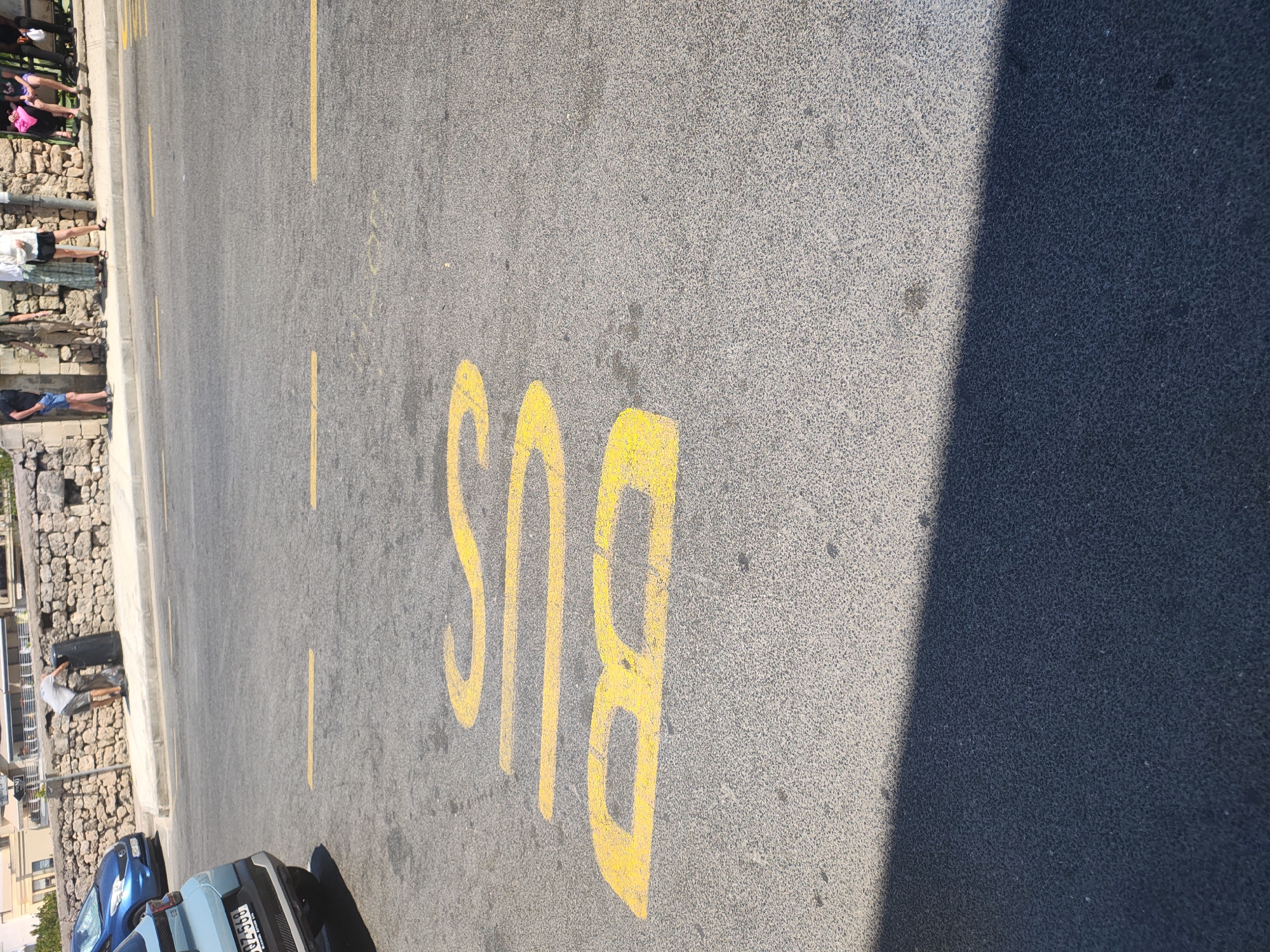 The image shows a street scene in Mellieħa, Malta. In the foreground, yellow painted letters spell out "BUS" on the asphalt. To the left, the rear of a light blue car is visible, parked on the side of the road. Behind the car, a blue sedan is also parked. In the background, a stone wall runs along the edge of the road, and several people are standing or sitting on the sidewalk. One person is seen near a black trash bin. The scene appears to be a bus stop area.