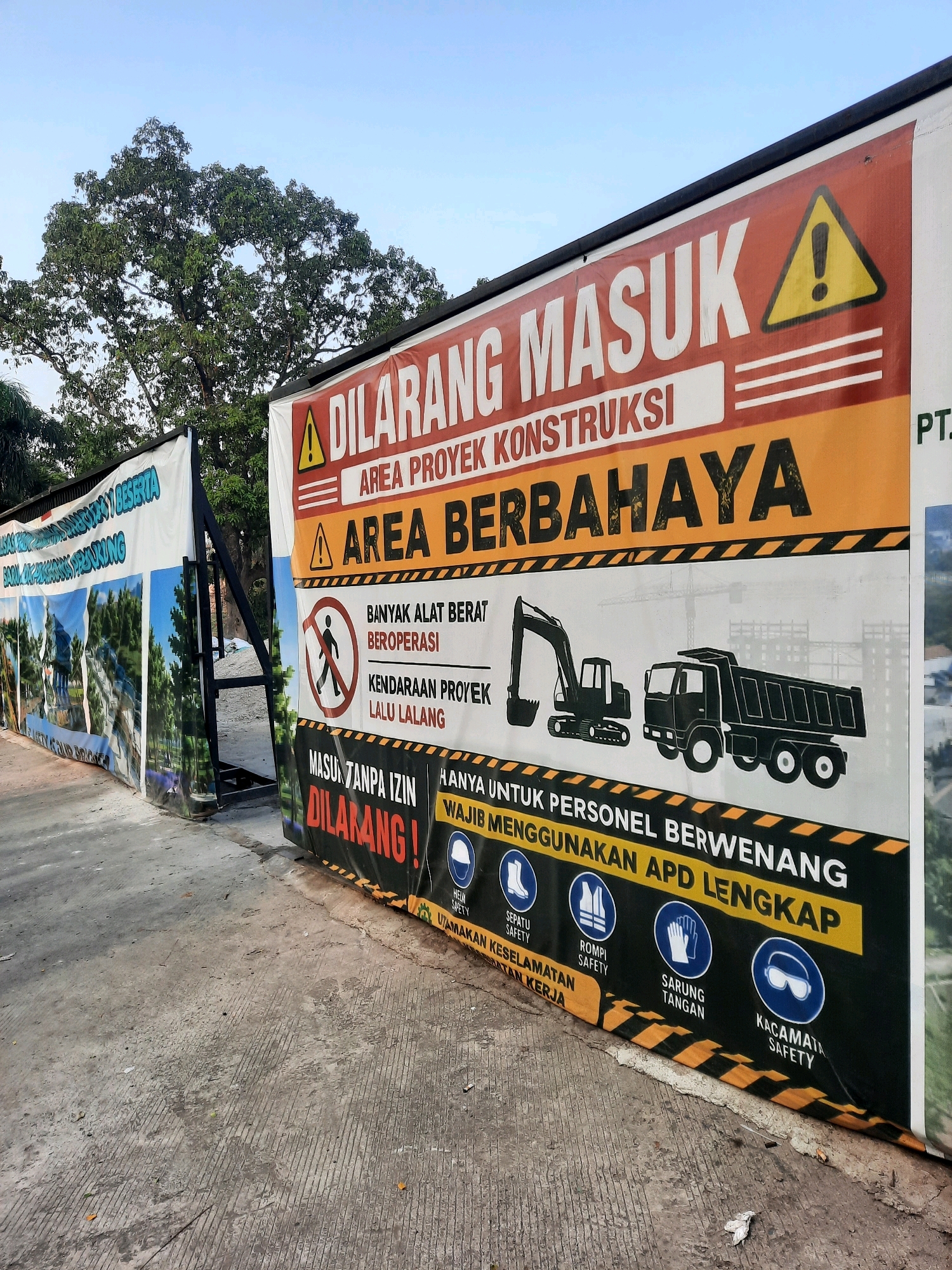 The image shows a construction site entrance in East Jakarta, Indonesia. A large banner displays "DILARANG MASUK" (No Entry) in red and white, with a yellow warning triangle. Below, it states "AREA PROYEK KONSTRUKSI" (Construction Project Area) and "AREA BERBAHAYA" (Dangerous Area). There are illustrations of an excavator and a dump truck, along with pictograms indicating safety equipment requirements such as boots, vests, gloves, and safety glasses. Another banner on the left, partially visible, shows images of buildings and green spaces, likely a representation of the completed project. The ground is paved with concrete, and a large tree stands in the background. The sky is clear and blue.