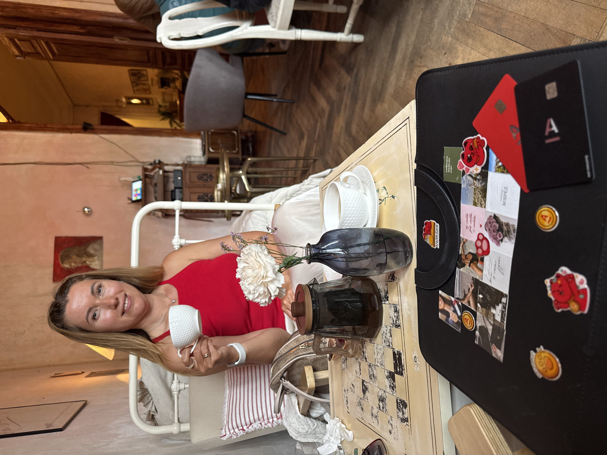 A woman with blonde hair and fair skin is sitting in a cafe, holding a white teacup in her right hand. She is wearing a red tank top and a white bracelet on her left wrist. In front of her is a tray with a glass teapot, a vase with a white flower and purple sprigs, and a cup and saucer. The tray is placed on a table with a checkered pattern. A black portfolio with various stickers and cards is in the foreground. The background shows a room with ornate wooden doors, a chair, and a piece of furniture with a tablet screen. The lighting is warm and inviting.
