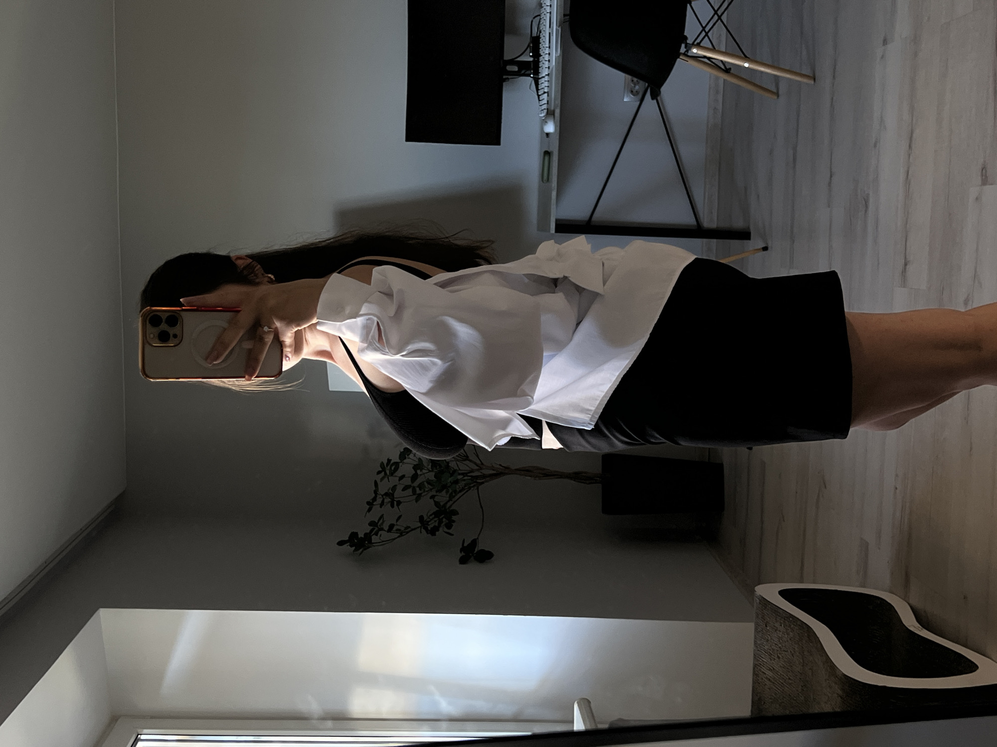 In a room in Ternopil, Ukraine, a woman is taking a mirror selfie. She is wearing a black top and a black skirt, with an unbuttoned white shirt worn over it. A smartphone is held up to obscure her face. The background includes a window with natural light, a potted plant, a desk with a computer monitor and keyboard, a chair, and a unique furniture piece in the foreground. The flooring is light-colored wood laminate.