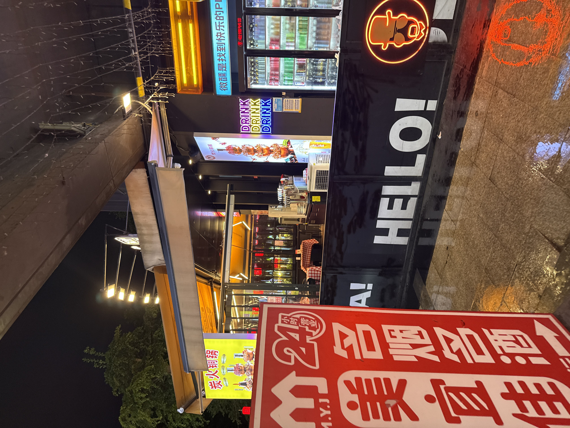 This is a nighttime photograph of a street-level storefront in Haizhu District, China. The scene is illuminated by various neon signs and interior lighting, suggesting it is likely evening or night. The overall atmosphere appears vibrant and possibly bustling, typical of a commercial area.
The foreground is dominated by a large red sign with white Chinese characters and an arrow pointing to the right. Visible text on this sign includes "24小时营业" which translates to "24-hour business," and "美宜佳" which is the name of a convenience store chain. Below this, the characters "名烟名酒" suggest that tobacco and alcohol are sold.
The main subject of the image is a food and beverage establishment, likely a restaurant or a convenience store with food offerings. A prominent black facade features large white lettering spelling "HELLO!" and an orange neon outline of a person wearing a hat and sunglasses. To the left, a bright yellow sign displays images of skewers, with Chinese characters "炭火铜锅" indicating a specialty like "charcoal hot pot." Nearby, neon signs with the word "DRINK" in colorful letters are visible. Behind the counter, a refrigerated display case is stocked with various beverages.
The setting appears to be a street or pedestrian walkway, with a tiled floor that appears wet, possibly from recent rain, reflecting the colorful lights. The architecture suggests a modern urban environment with some decorative string lights overhead. No people are clearly visible in the photograph, but the setting implies a place of commerce and activity. The neon lights and signage contribute to a lively and commercial ambiance.