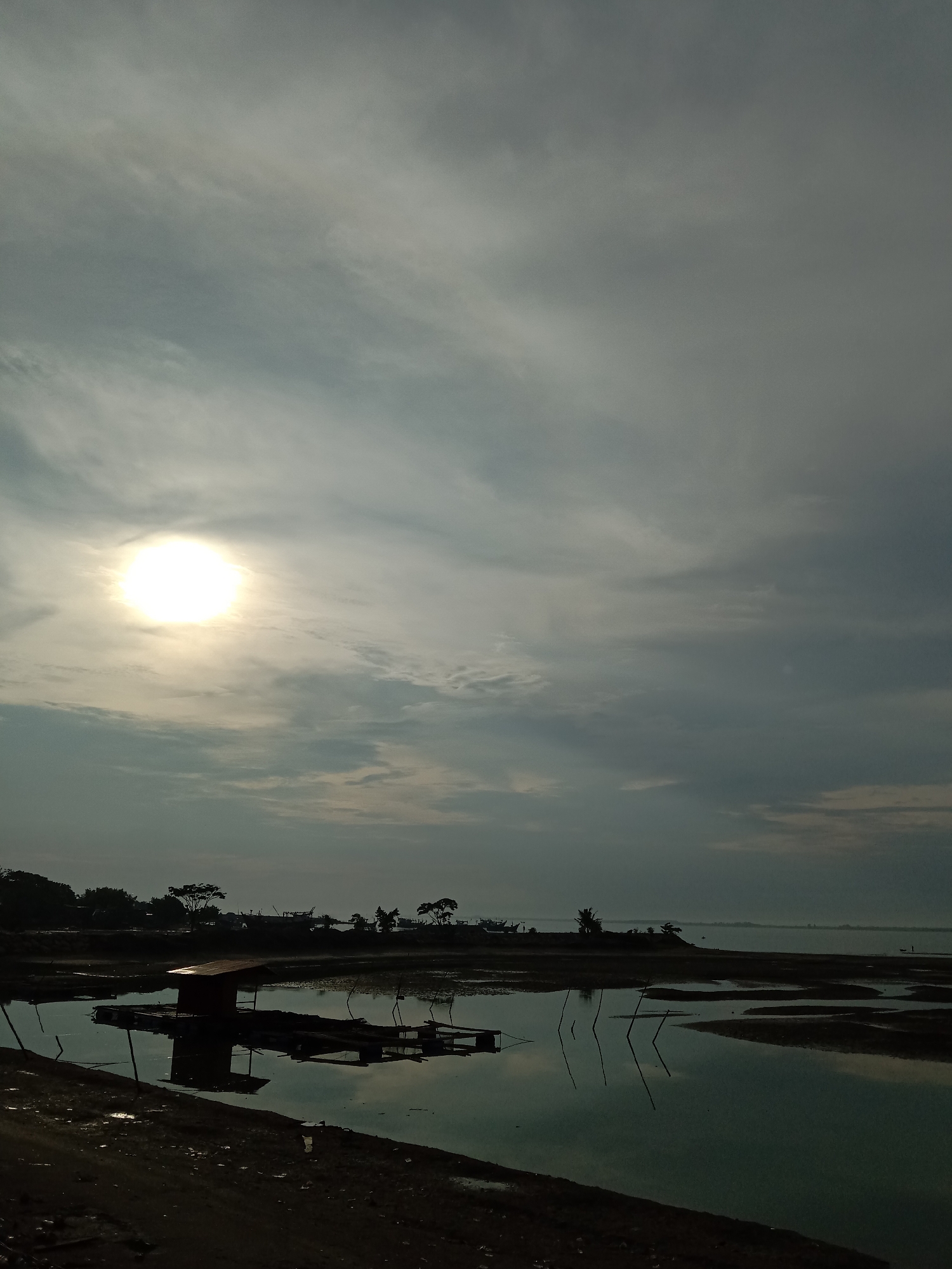 The image depicts a tranquil scene on a body of water under an overcast sky, likely in Lhokseumawe, Indonesia. The sun is visible through the clouds, casting a diffused light. In the foreground, shallow water reflects a small, wooden structure, possibly a raft or a shelter, and the surrounding landscape. Several wooden poles are also visible, seemingly embedded in the water and reflecting in its surface. The surrounding land appears muddy or sandy. In the midground, the shoreline is marked by silhouettes of trees and other structures. The horizon appears somewhat obscured by haze.