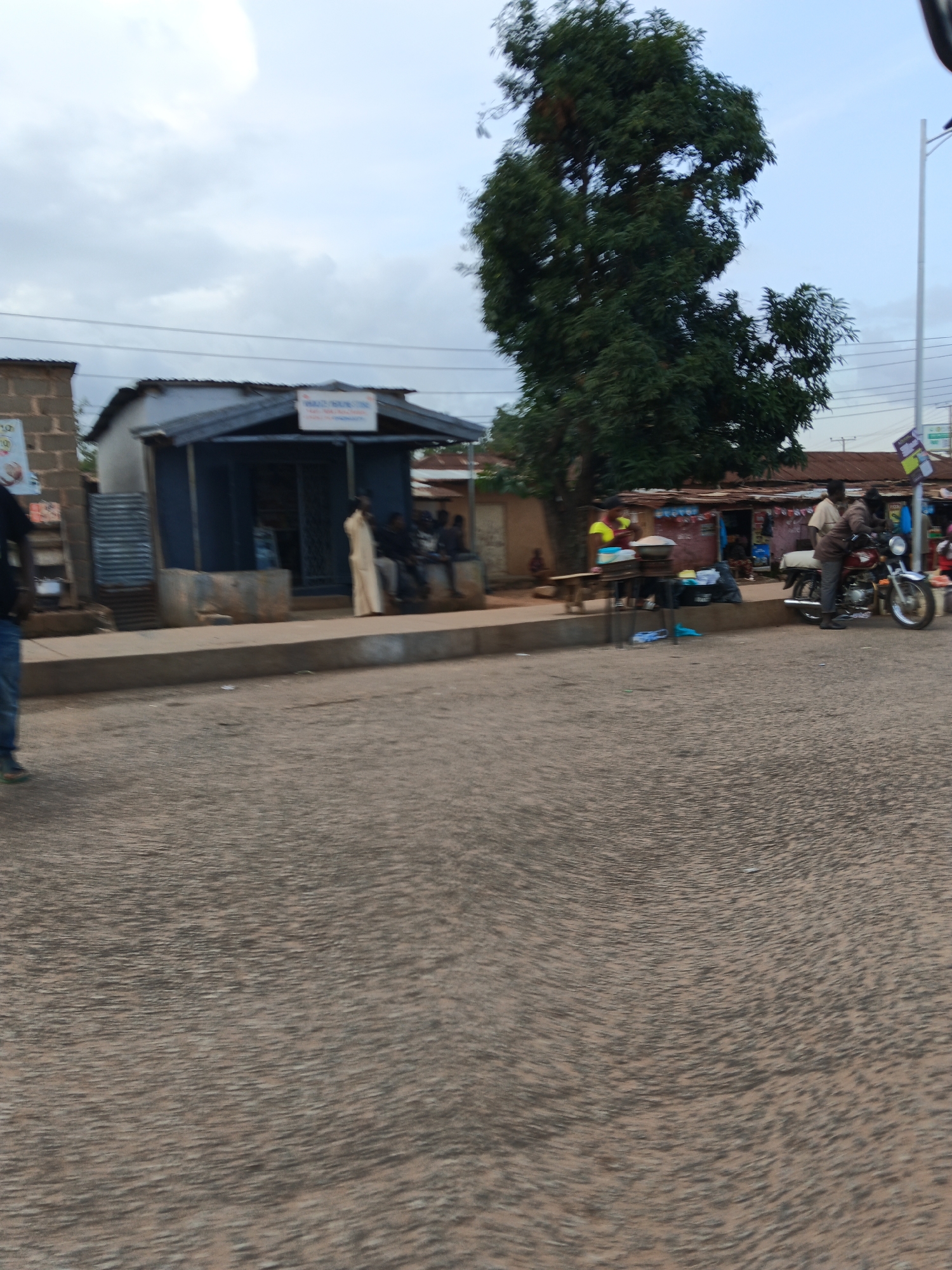 This image depicts a street scene in Bukuru, Nigeria, under a cloudy sky, suggesting it's either early morning or late afternoon. The foreground is dominated by a textured asphalt road.
In the mid-ground, a paved walkway runs parallel to the road, bordered by a low concrete barrier. Behind the barrier are several small shops and structures. On the left, a blue building with a metal roof and a sign that appears to read "MOBILE PHONE" is visible. People are gathered in front of and inside this building, some sitting on a bench. To the right of this blue shop, there's a market-like area with makeshift stalls covered by corrugated metal roofs. A large tree with dense foliage stands prominently behind these stalls, casting some shade.
Several people are engaged in various activities. On the right side of the market area, two individuals are interacting with a red motorcycle. One person is standing next to the motorcycle, appearing to be adjusting something on it, while another is seated on it. Further to the left, near the blue shop, a person in a light-colored garment stands outside. Other individuals are seated on a ledge in front of the shop. A person in blue jeans and a black shirt is visible on the far left of the image, walking away from the camera.
The overall scene suggests a typical day in a bustling local market or commercial area. The presence of shops, people congregating, and a motorcycle points to a place of commerce and daily life. The cloudy weather contributes to a diffused light, and the environment seems to be an outdoor urban setting.