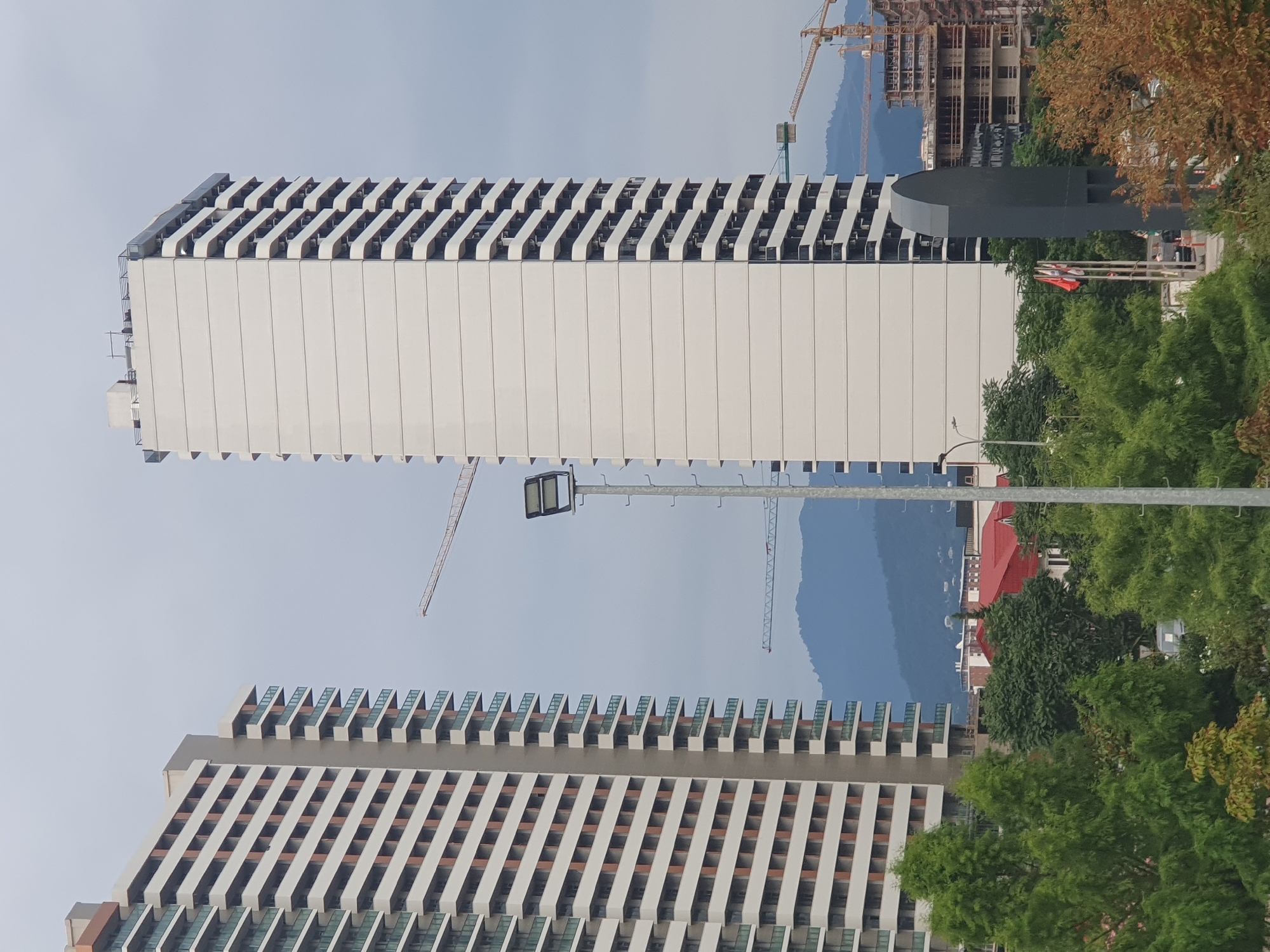 The image captures a cityscape in Batumi, Georgia, dominated by two large, modern apartment buildings. The building on the left is tall and rectangular with a facade characterized by alternating white and brown horizontal bands, interspersed with balconies. To its right, a taller, slender white building features a repeating pattern of balconies that curve inwards. In the background, a range of blue mountains forms a natural backdrop against an overcast sky.
Several construction cranes are visible, indicating ongoing development in the area. One crane is positioned near the top left of the image, extending across the sky. Another, smaller crane is visible between the two apartment buildings, and a larger construction crane is situated on the right side of the frame, near a partially constructed building with visible scaffolding.
In the foreground, lush green trees and foliage obscure the lower part of the scene. A tall, slender street lamp with a rectangular light fixture stands prominently in the center-right foreground. To its left, a dark, curved architectural structure is present, and behind it, a flagpole with several flags can be seen. Further to the right, the skeletal structure of a building under construction is evident, with cranes actively working around it. The overall impression is one of urban development and construction set against a natural mountainous landscape.